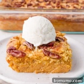 A square slice of pumpkin dump cake topped with pecans and a scoop of vanilla ice cream on a plate, with a baking dish of the dessert in the background.