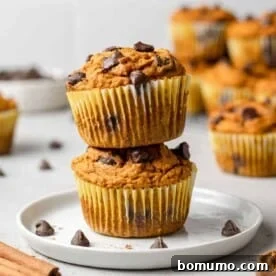 Two perfectly baked pumpkin chocolate chip muffins stacked on a small white plate, with more delicious muffins blurred in the background.