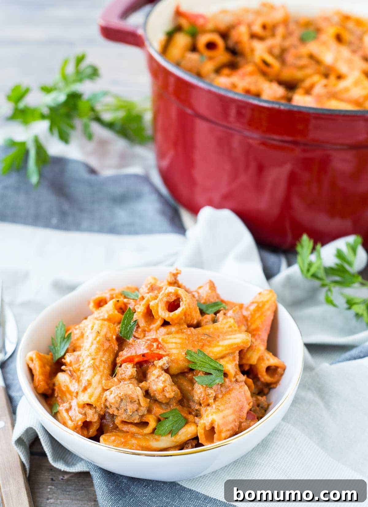 Small bowl of rigatoni pasta garnished with fresh parsley, with a vibrant red Dutch oven visible in the background.