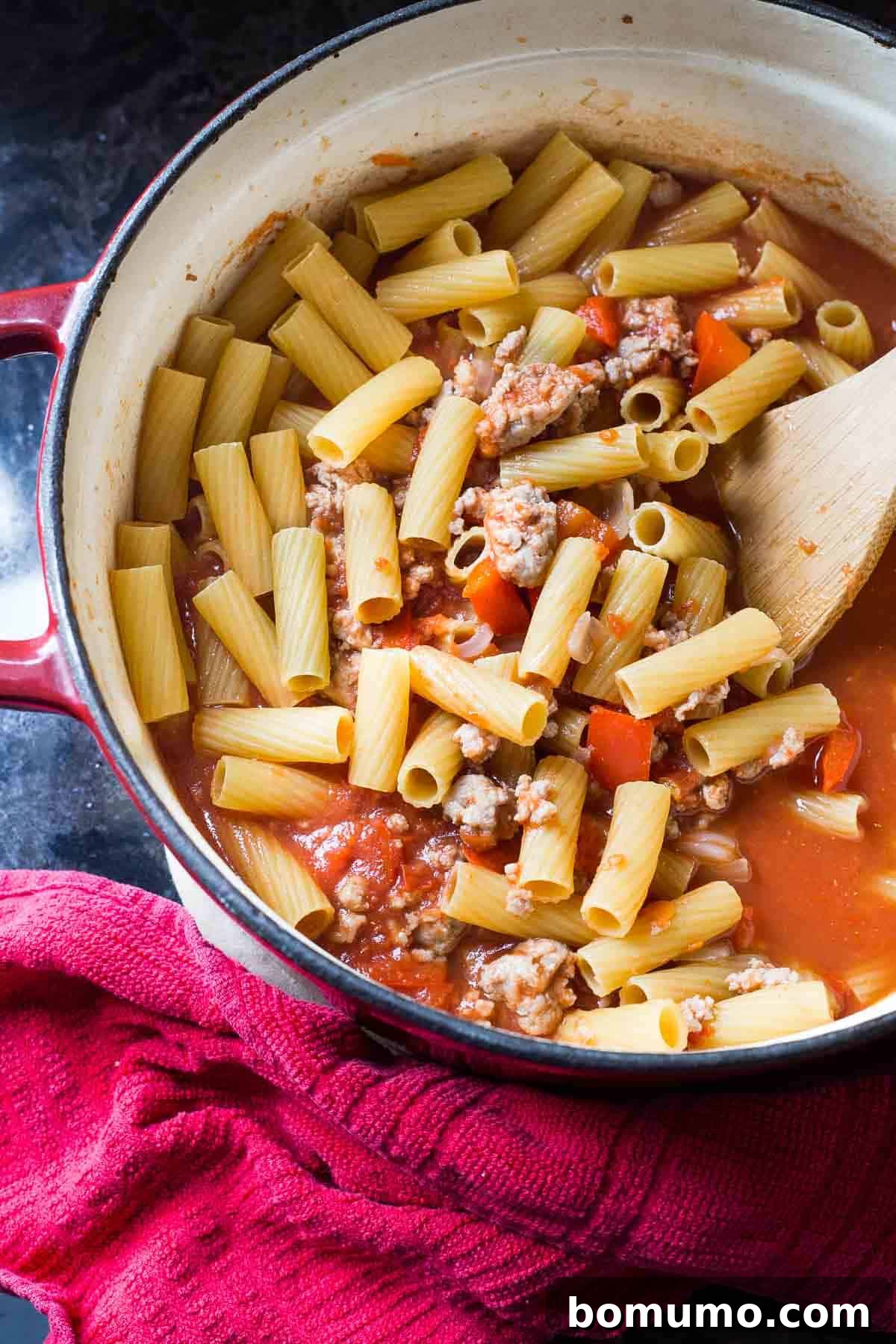 Overhead shot of a red Dutch oven containing uncooked rigatoni pasta, Italian sausage, and tomatoes, prior to cooking.
