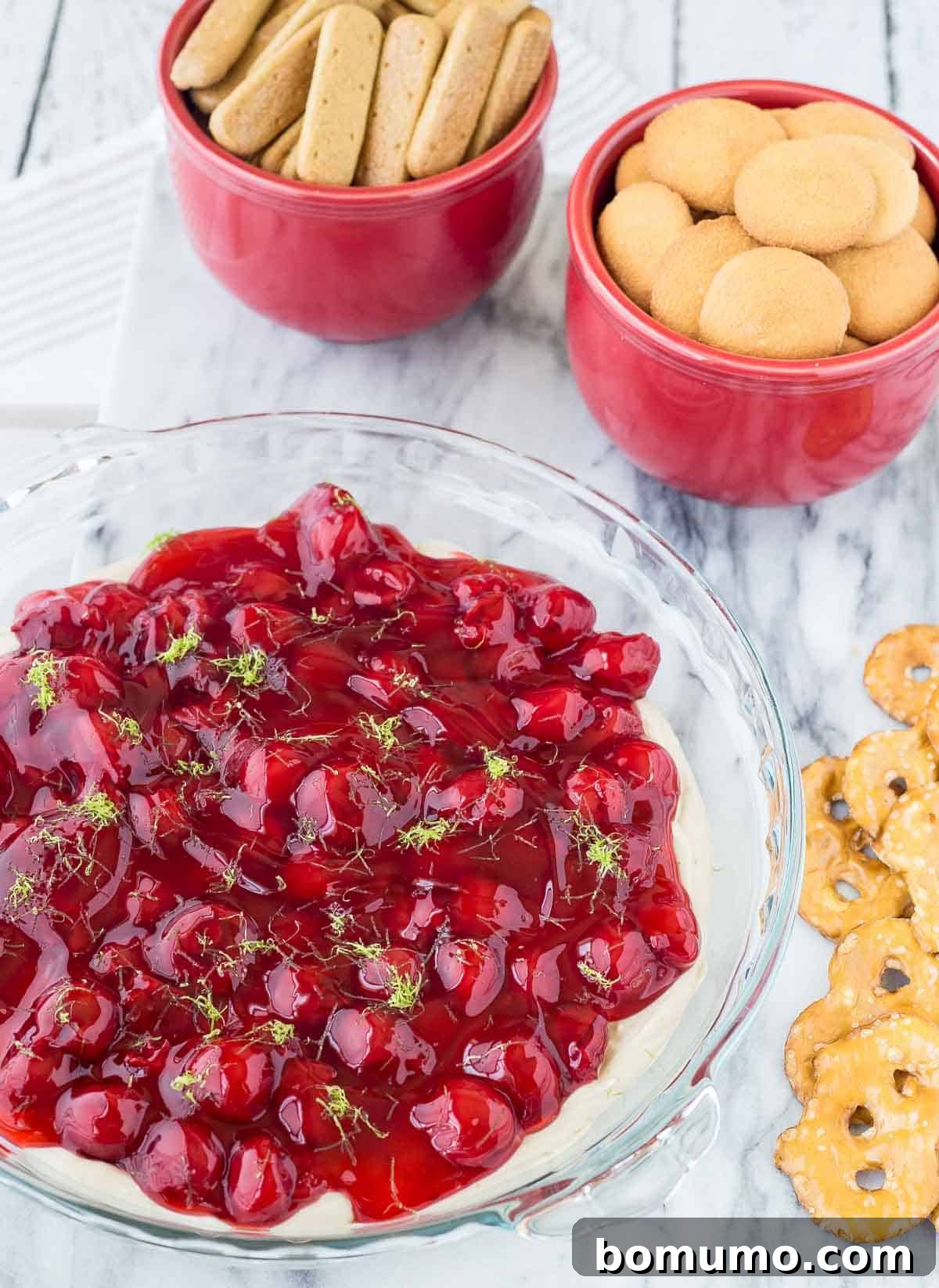 A vibrant cherry cheesecake dip in a pie pan, surrounded by various dippers like graham crackers and pretzels, showcasing its appealing texture and rich color.