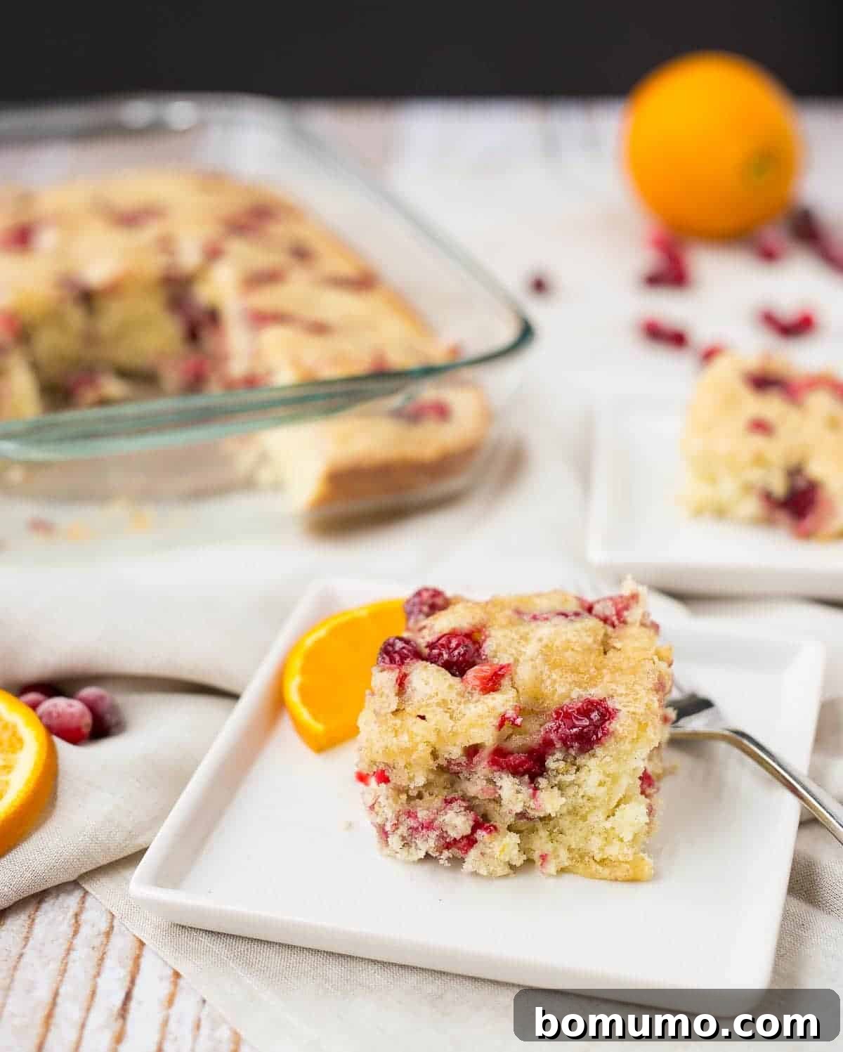 A slice of vibrant cranberry coffee cake on a square white plate with a fork, elegantly garnished with a half-moon orange slice.