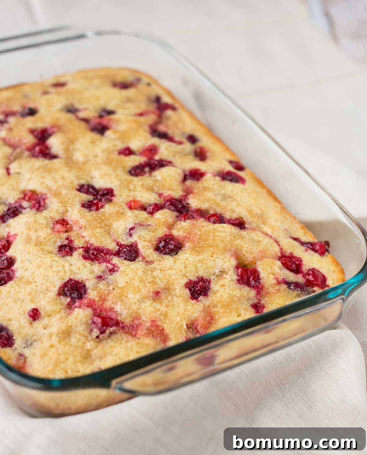 A partial overhead view of the cranberry coffee cake, freshly baked in a clear glass baking dish, resting on a white cloth background.