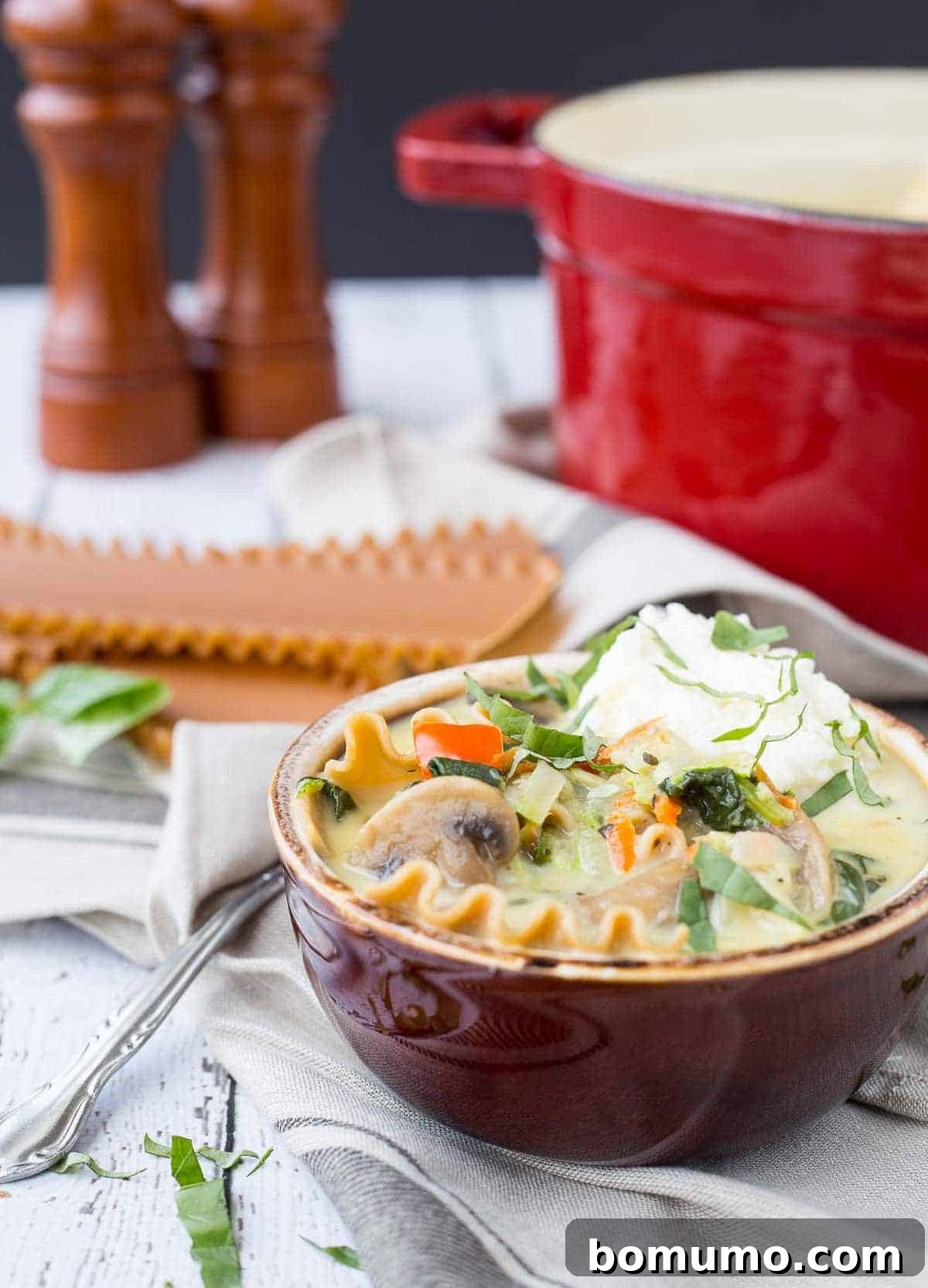A comforting serving of vegetarian lasagna soup in a rustic brown earthenware bowl, elegantly garnished with a dollop of creamy ricotta cheese and vibrant fresh basil leaves.