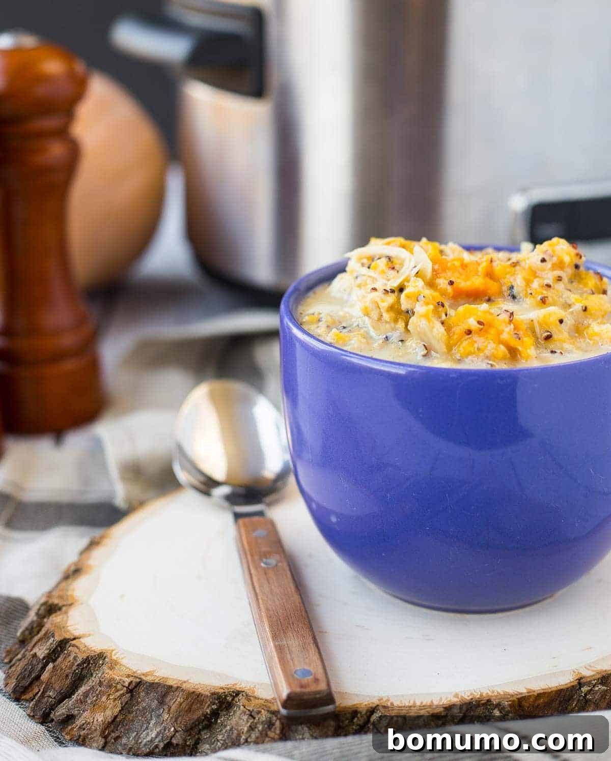 Front view of slow cooker creamy chicken soup in a rustic blue mug with a spoon, set on a wooden slice, evoking cozy fall vibes.