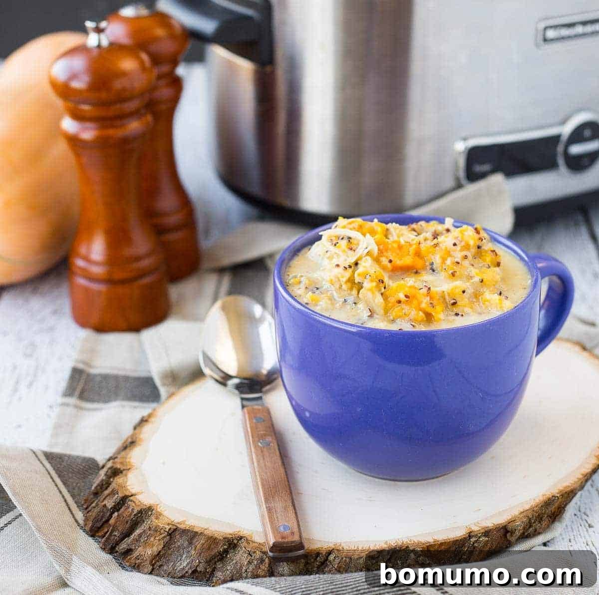 Slow cooker creamy chicken soup in a blue mug, with a slow cooker, butternut squash, and salt/pepper mills in the background, illustrating the wholesome preparation.