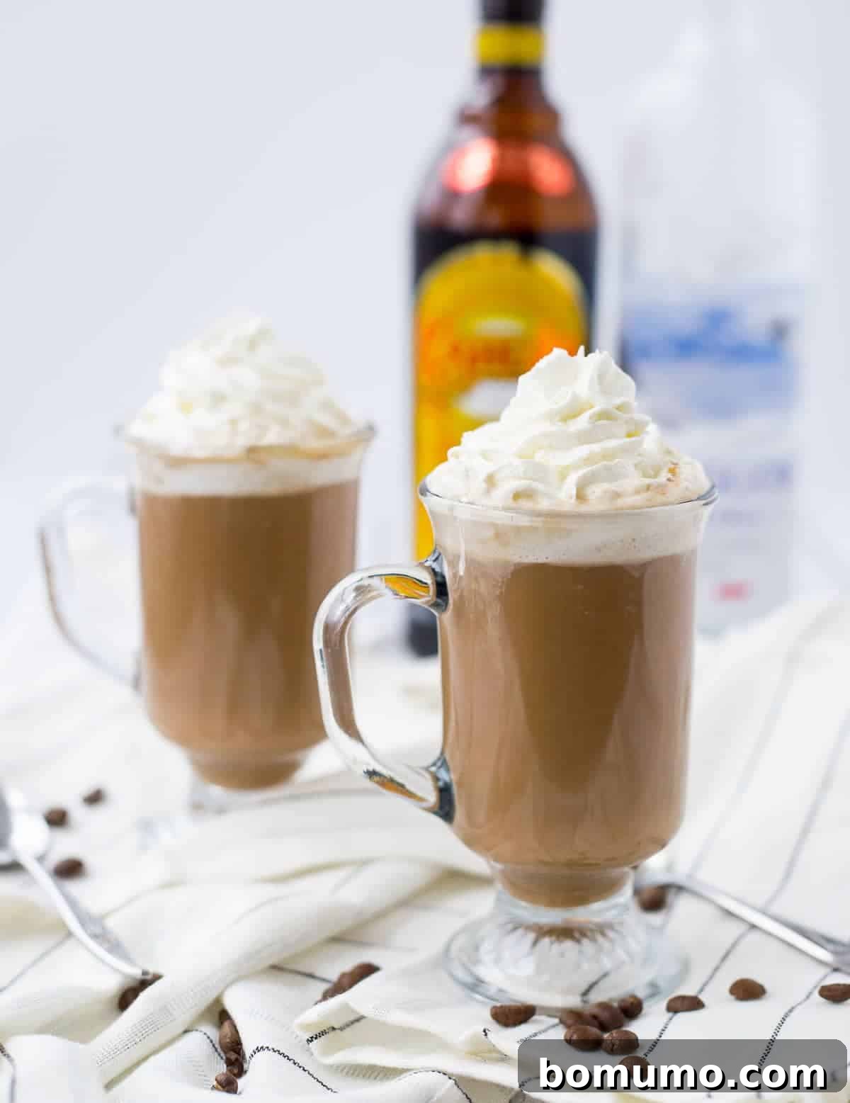Footed clear glass mugs containing hot chocolate garnished with whipped cream, on white striped cloth, with bottle of Kahlua in background.