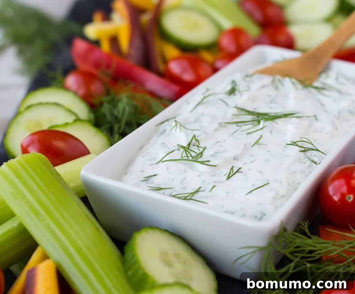 A white bowl filled with creamy dill vegetable dip, elegantly garnished with a sprig of fresh dill, surrounded by an assortment of colorful cut vegetables like cucumber slices, carrot sticks, and broccoli florets.