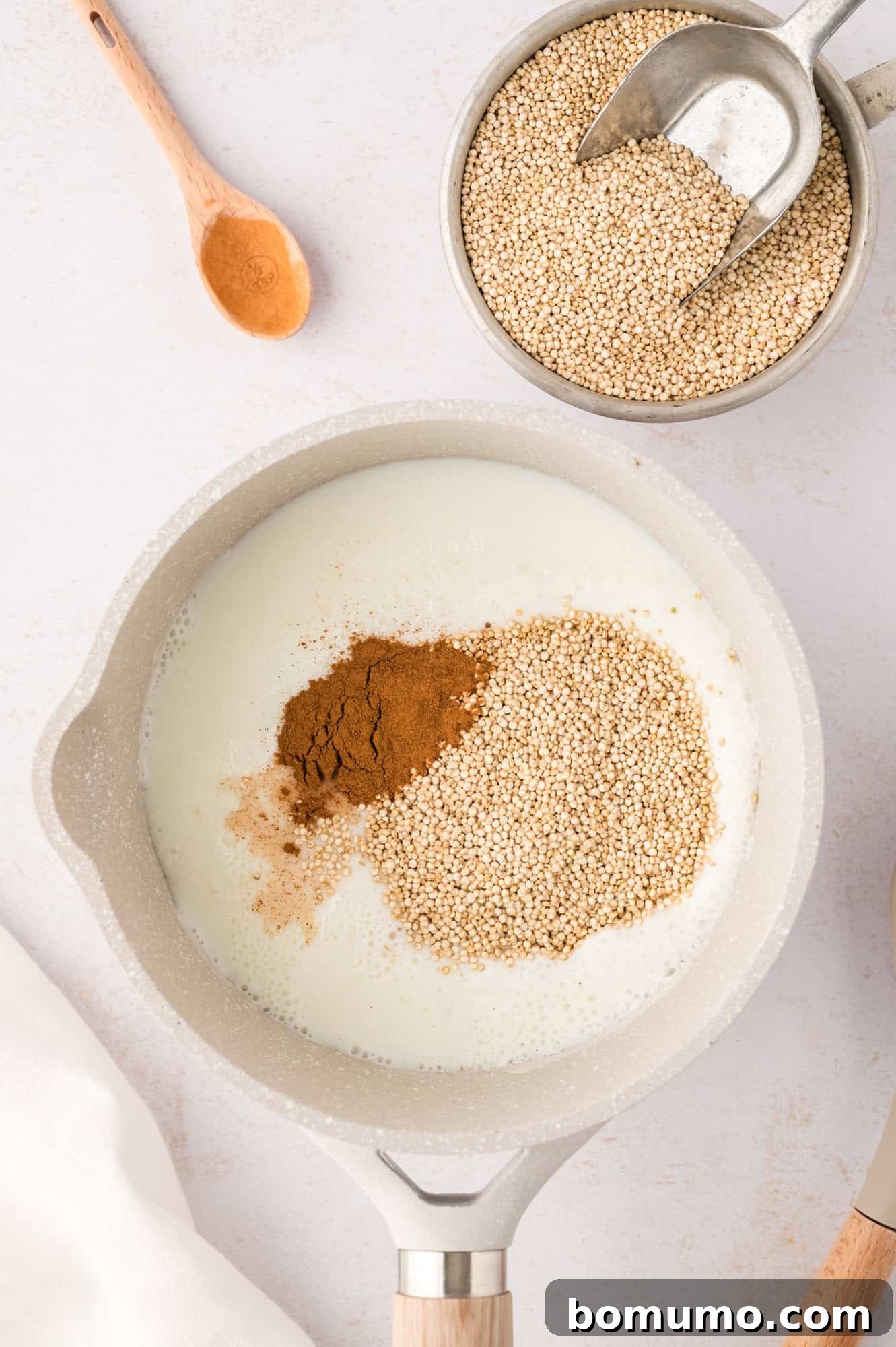 Rinsed quinoa grains and a sprinkle of ground cinnamon being carefully added to the simmering milk and water mixture in a saucepan.