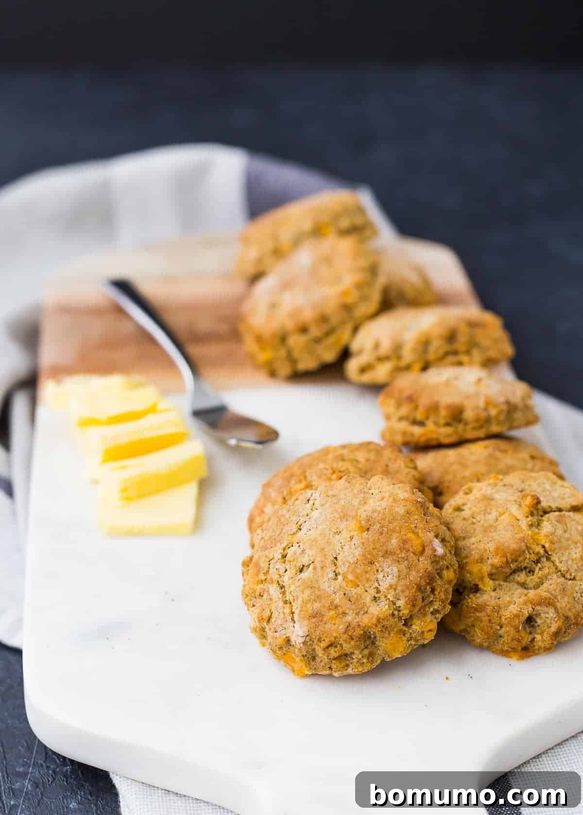 Several golden-brown whole wheat biscuits stacked on a white cutting board, accompanied by pats of butter and a spreader, ready to be enjoyed.