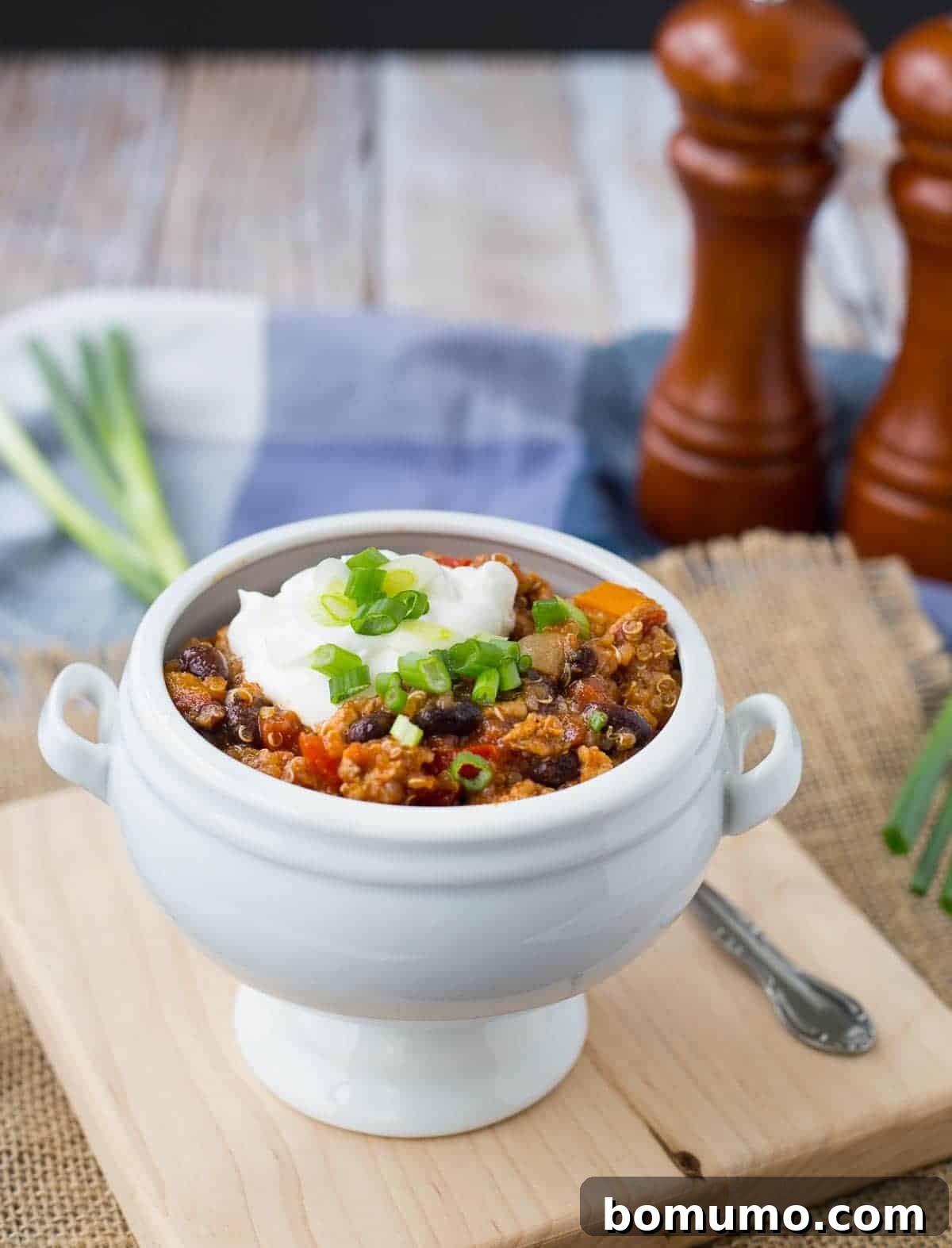 A beautifully garnished serving of turkey quinoa chili in a white bowl, topped with a dollop of sour cream and fresh green onions.