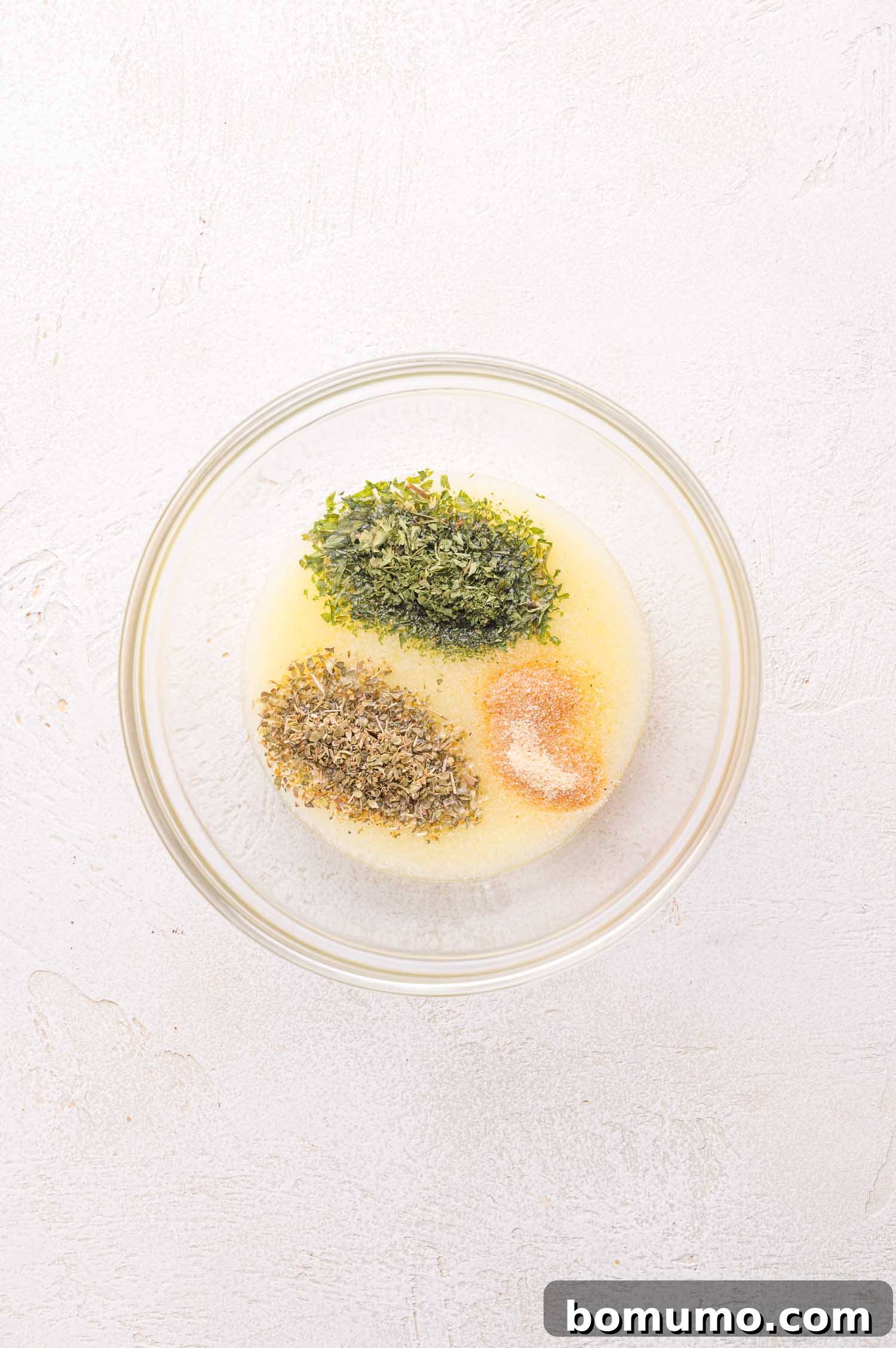 Gooey Pizza Pull-Aparts 11 A glass bowl containing melted butter, dried parsley, dried oregano, garlic powder, and salt on a light-colored surface.