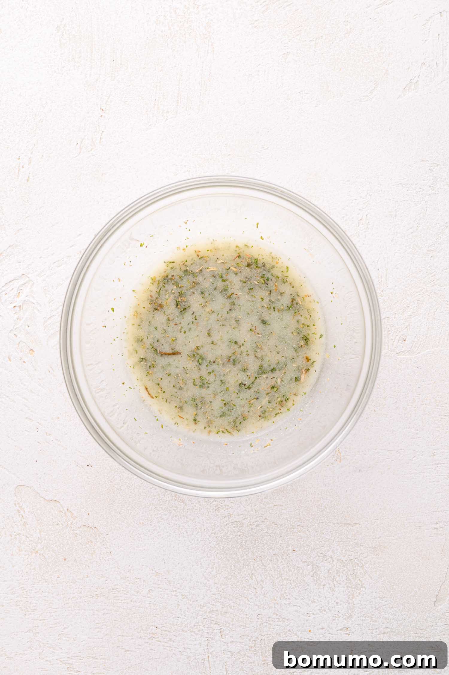 Gooey Pizza Pull-Aparts 12 A clear glass bowl containing a light-colored liquid mixed with green and brown dried herbs, placed on a light textured surface.