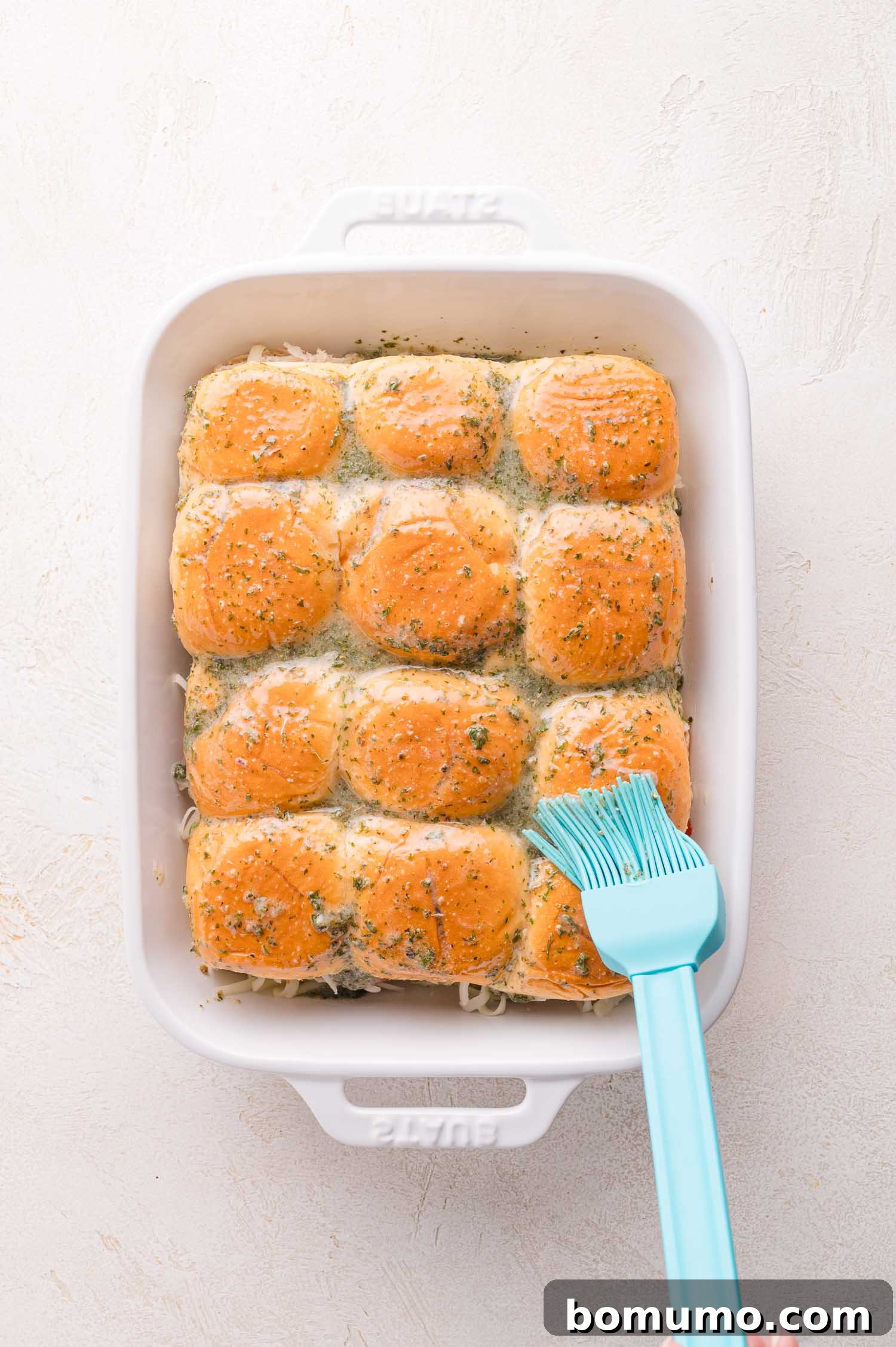 Gooey Pizza Pull-Aparts 14 A white baking dish with twelve golden-brown dinner rolls, some topped with herbs, being brushed with melted butter using a blue silicone brush.