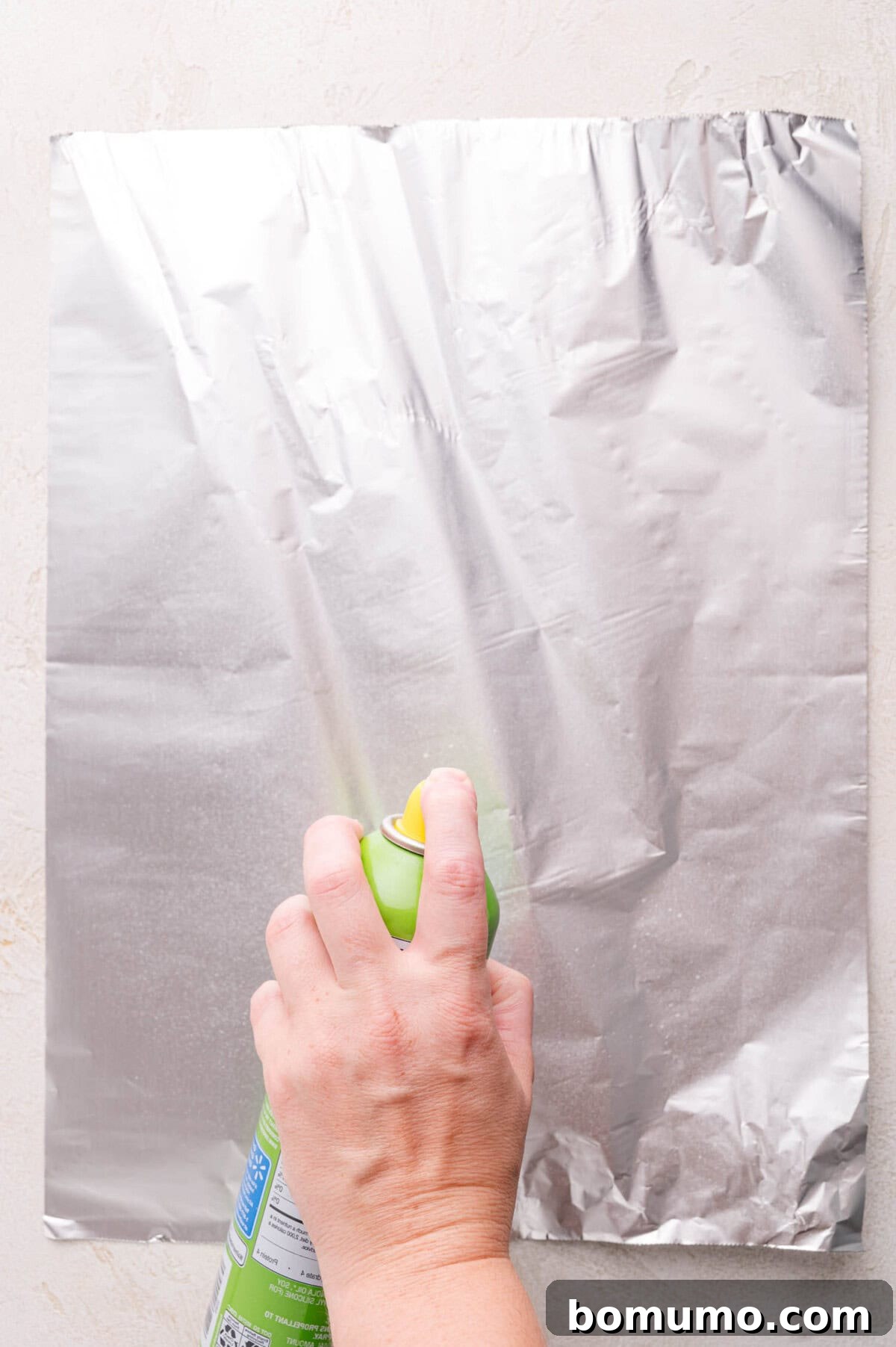 Gooey Pizza Pull-Aparts 15 A hand sprays cooking oil from a green can onto a sheet of aluminum foil placed on a light-colored surface.