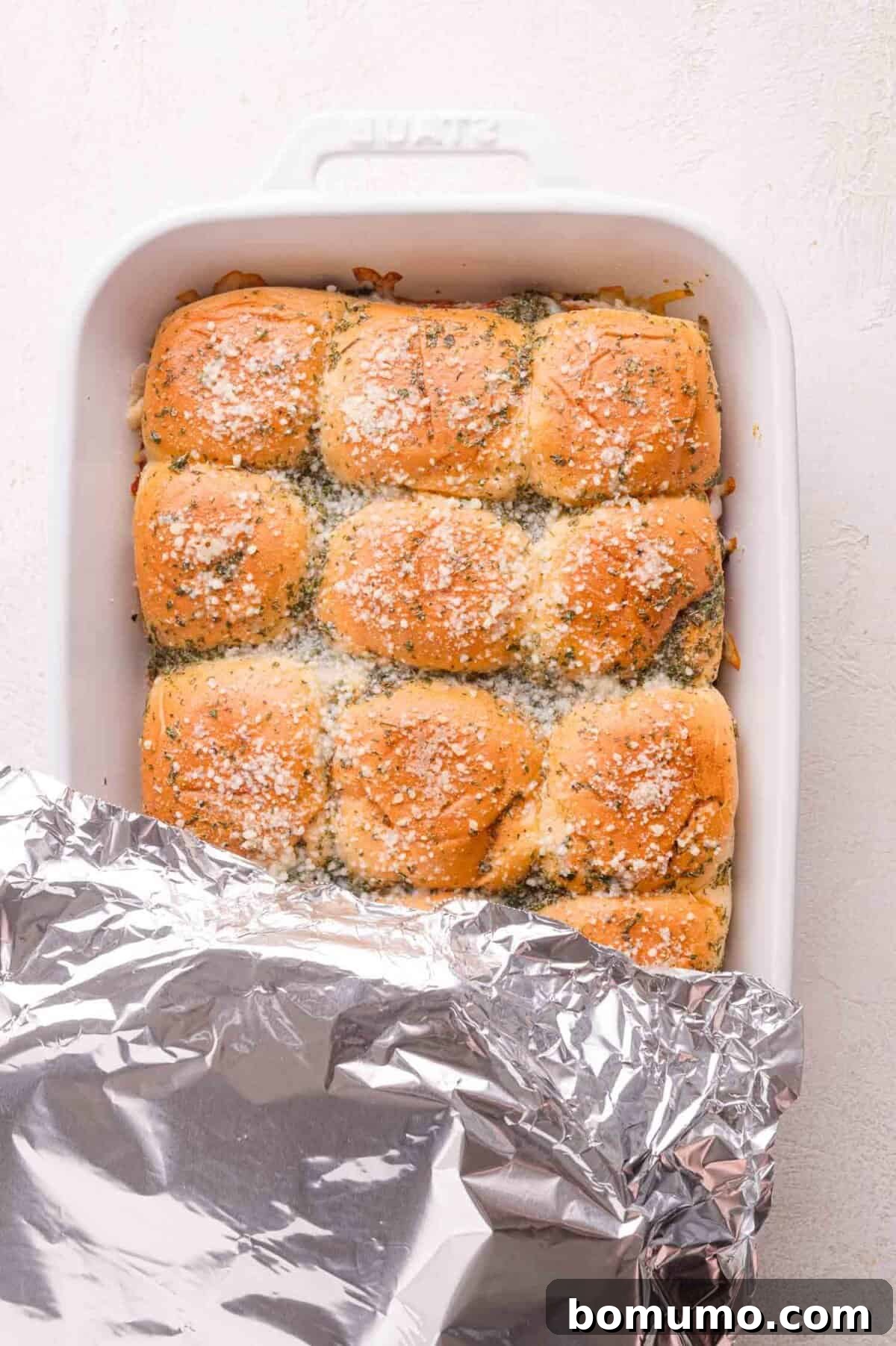 Gooey Pizza Pull-Aparts 16 A baking dish filled with golden brown dinner rolls topped with grated cheese and herbs, partially covered with aluminum foil.