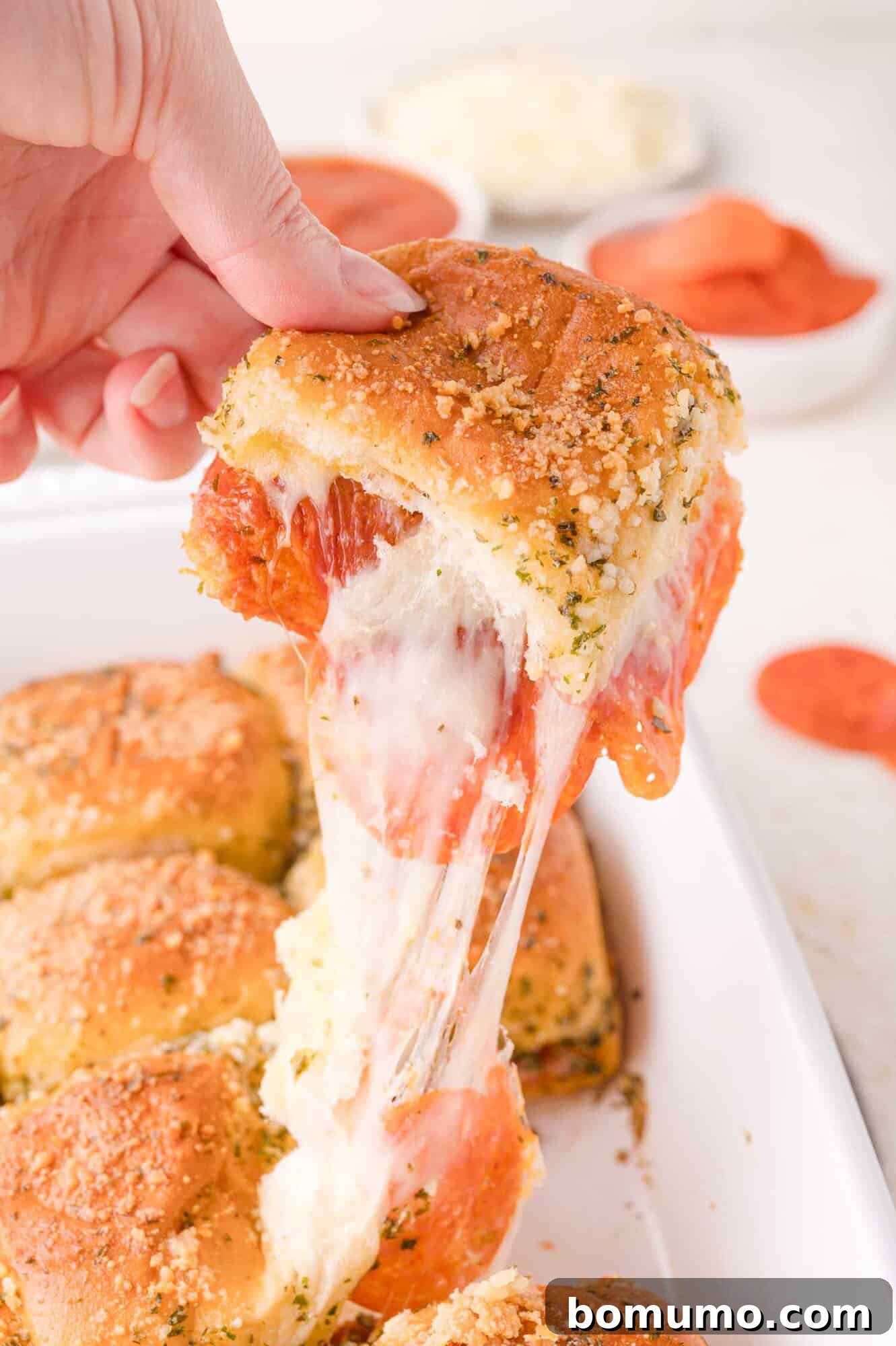 Gooey Pizza Pull-Aparts 17 A hand lifts a cheesy pepperoni slider from a baking dish, showing melted cheese stretching from the sandwich.