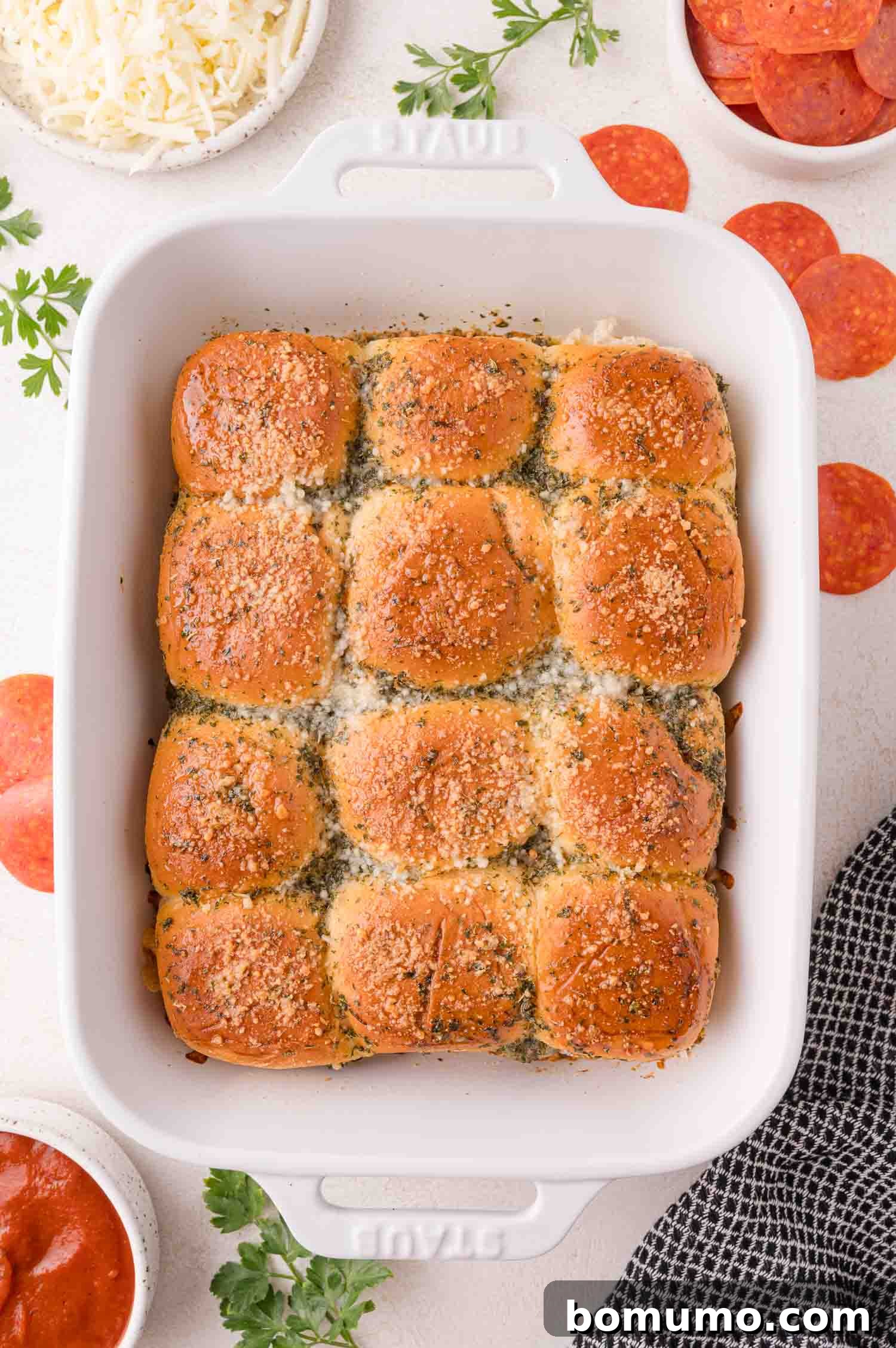 Gooey Pizza Pull-Aparts 3 A white baking dish filled with twelve golden-brown slider buns topped with herbs and grated cheese, surrounded by small bowls of shredded cheese, pepperoni, and sauce.