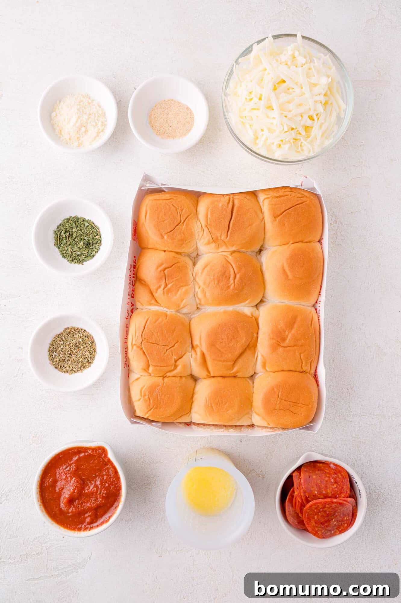 Gooey Pizza Pull-Aparts 4 Top-down view of slider buns in a tray surrounded by bowls of shredded cheese, pepperoni slices, marinara sauce, melted butter, and seasonings on a white surface.