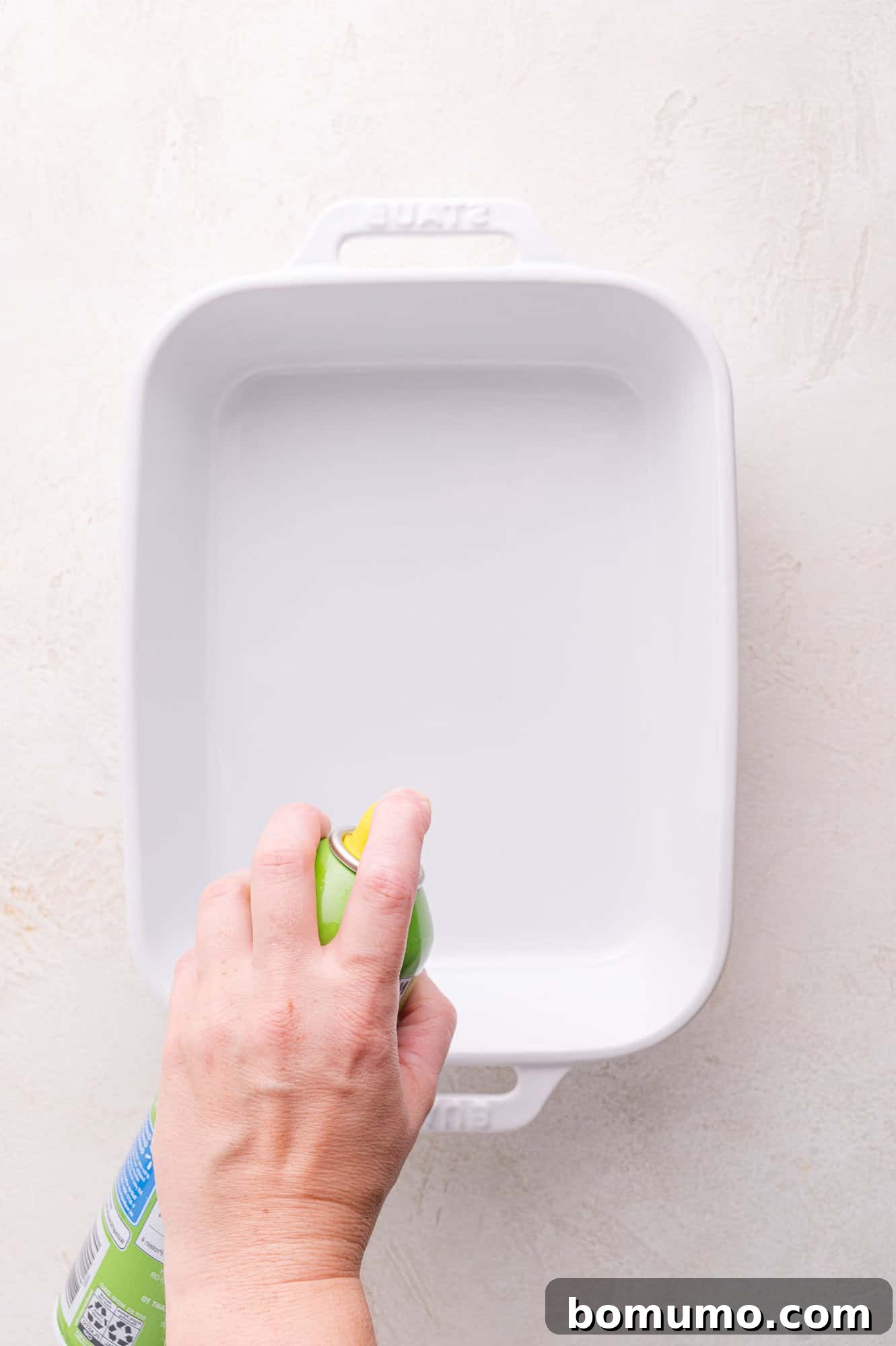 Gooey Pizza Pull-Aparts 5 A hand sprays nonstick cooking spray onto a white rectangular baking dish placed on a light-colored surface.