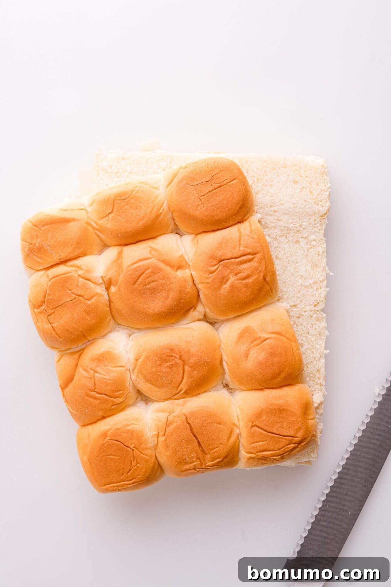 Gooey Pizza Pull-Aparts 6 A set of twelve slider buns partially sliced in half on a white surface, next to a serrated knife.