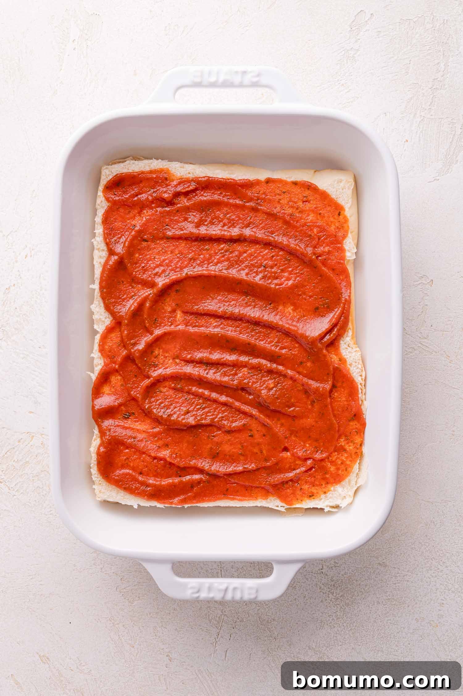 Gooey Pizza Pull-Aparts 7 A white baking dish with a single layer of lasagna noodles topped with tomato sauce on a light-colored surface.