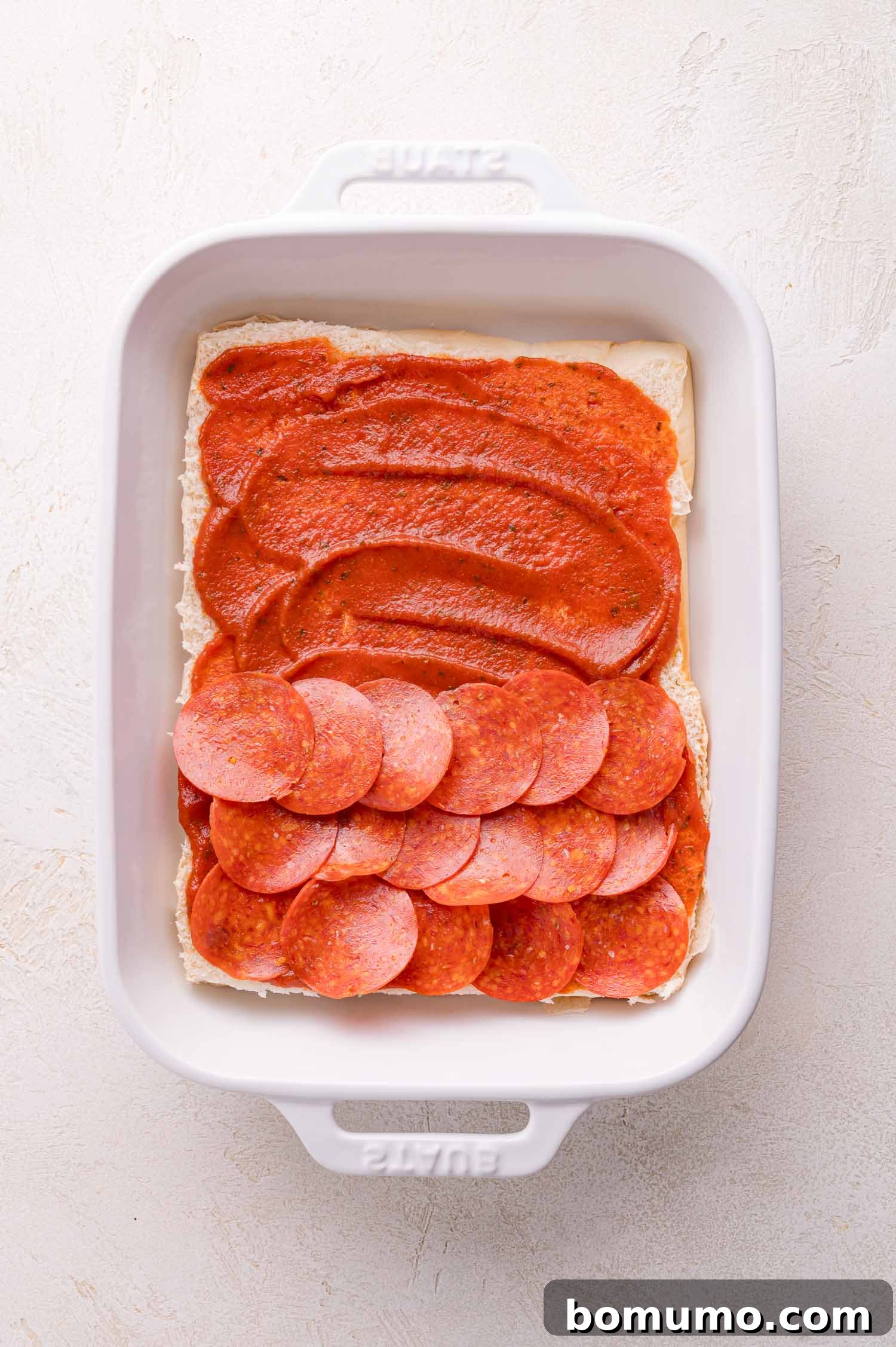 Gooey Pizza Pull-Aparts 8 Rectangular white baking dish containing a layer of flat dough, spread with tomato sauce, and topped with slices of pepperoni on one half.