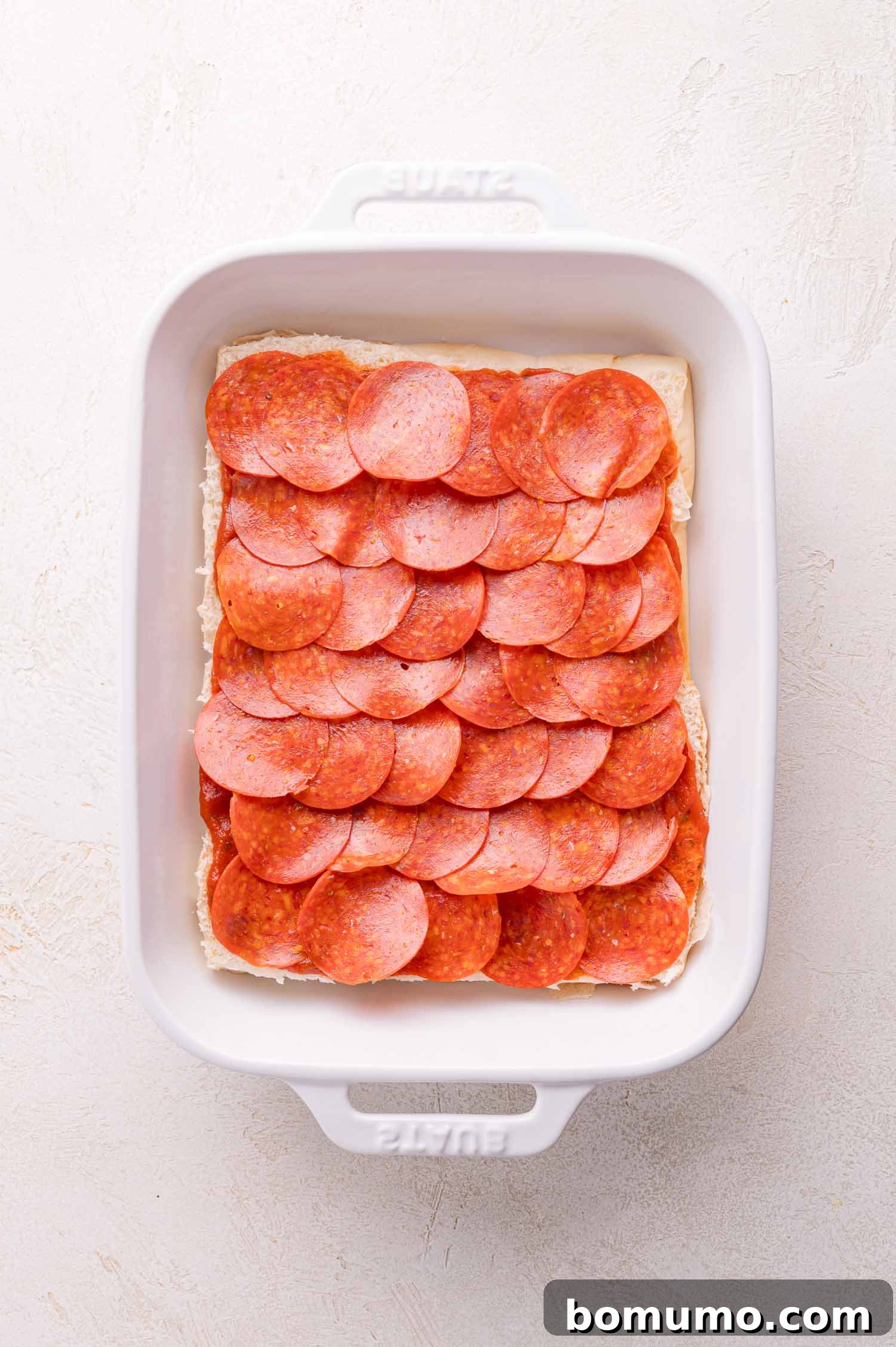Gooey Pizza Pull-Aparts 9 A white baking dish with a layer of overlapping pepperoni slices arranged on top of an uncooked rectangular base.