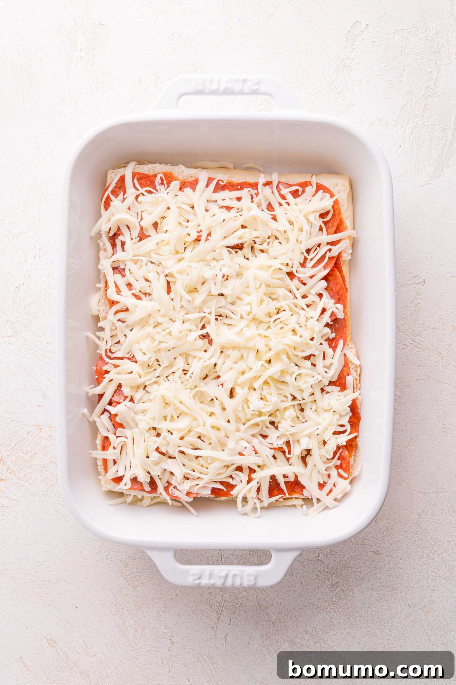 Gooey Pizza Pull-Aparts 10 A white baking dish with an unbaked layer of lasagna topped with tomato sauce and shredded cheese.