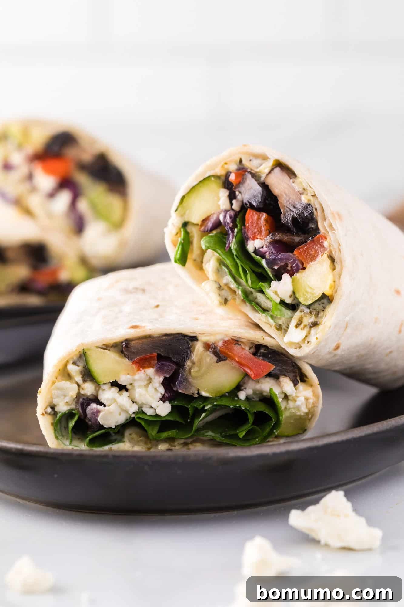 Two halves of a vibrant roasted vegetable wrap, revealing tender vegetables, feta, and spinach, presented on a sleek black ceramic plate.