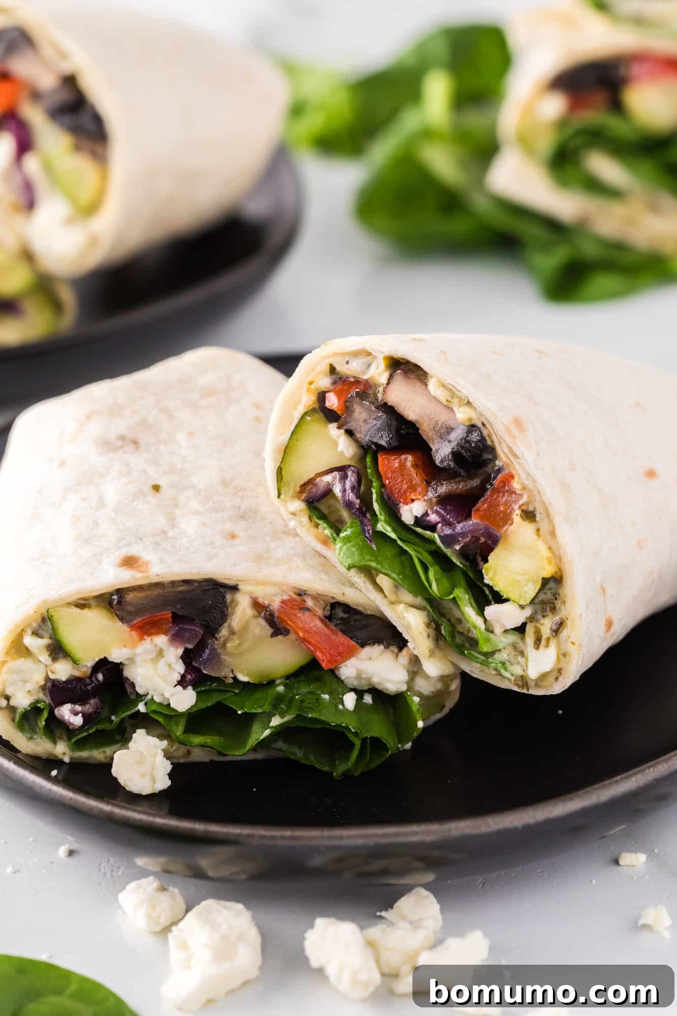 Two perfectly cut halves of a roasted vegetable wrap, filled with colorful veggies, feta, and spinach, presented on a black ceramic plate.
