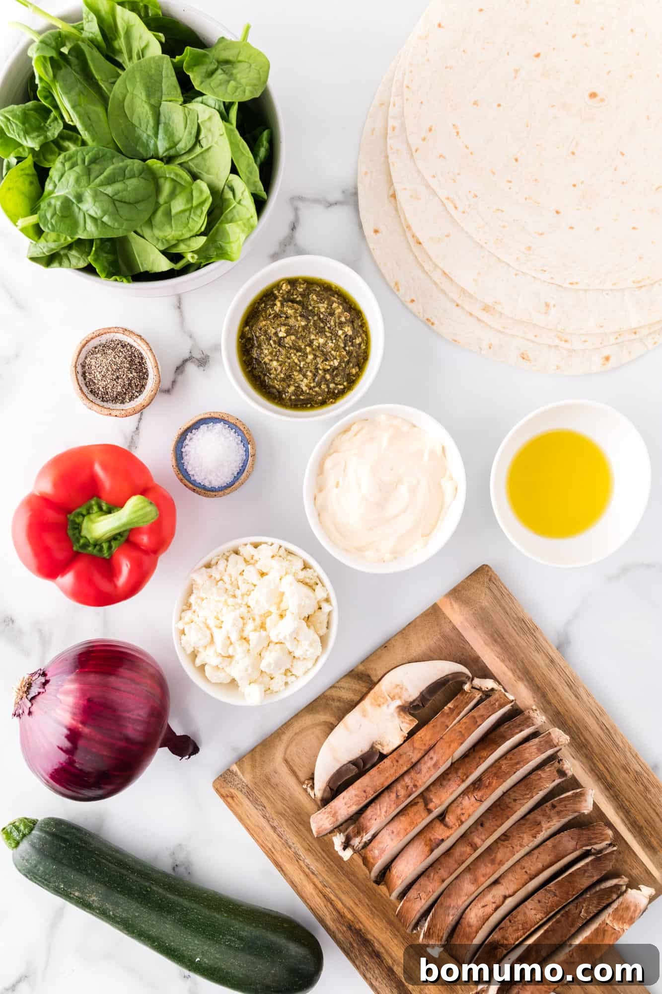 A vibrant collection of fresh ingredients laid out for making roasted vegetable wraps, including bell peppers, zucchini, mushrooms, onions, spinach, feta, and wraps.