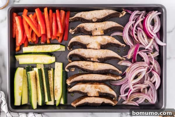 Precisely sliced zucchini, bell peppers, onions, and mushrooms arranged on a baking sheet, ready for seasoning and roasting.