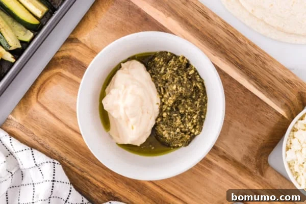 Mayonnaise and vibrant green pesto being combined in a small white bowl, resting on a wooden cutting board.
