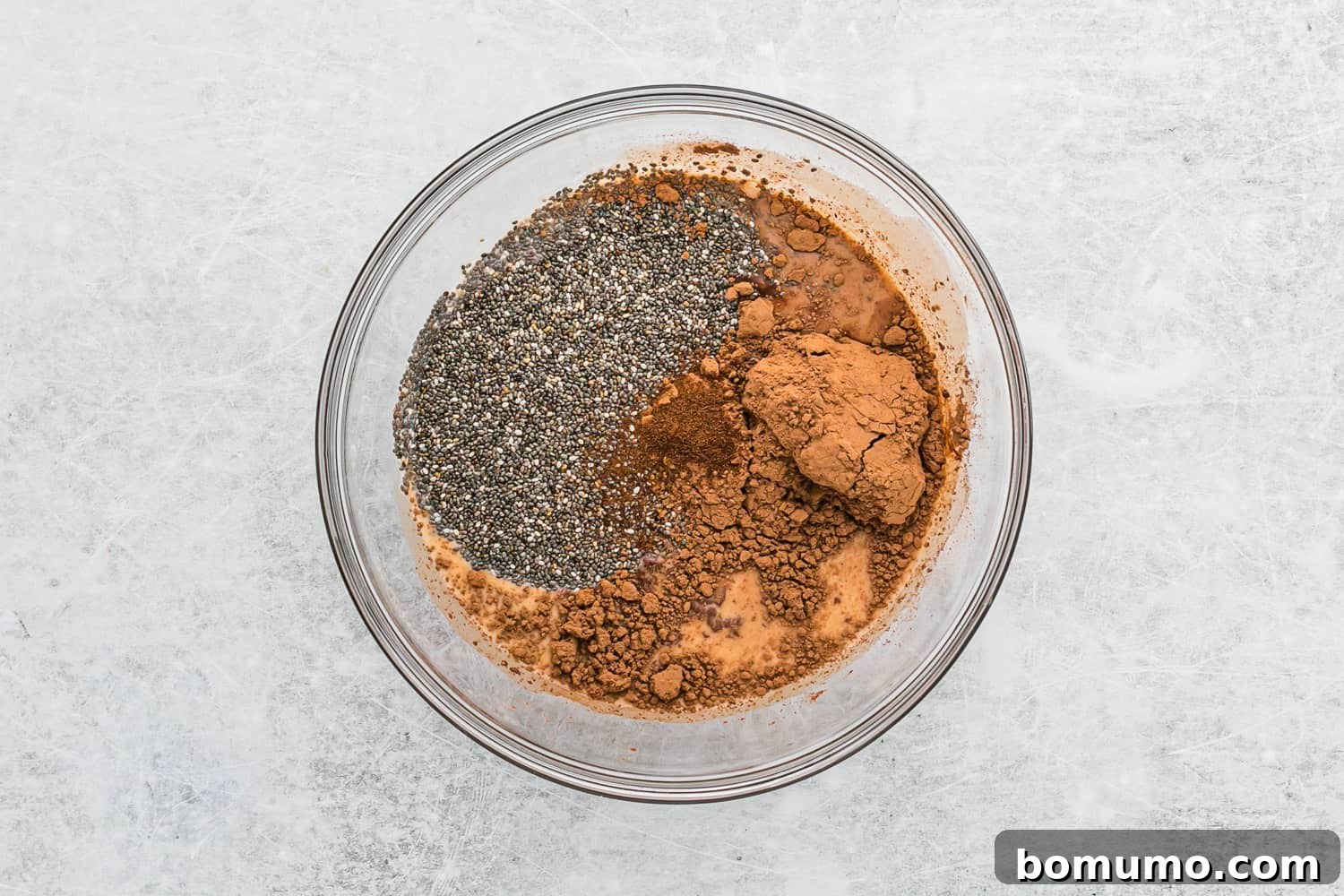 Unmixed ingredients for chocolate chia pudding, including milk, chia seeds, cocoa, maple syrup, and vanilla, resting in a clear glass bowl.