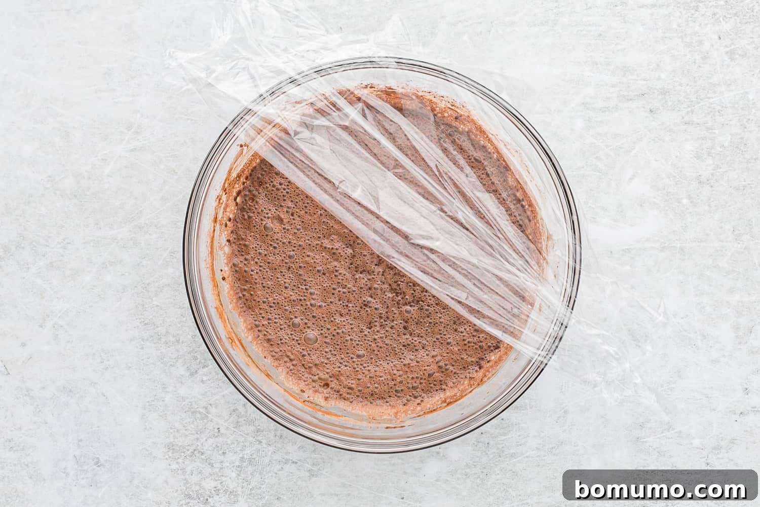 A glass bowl containing the chocolate chia pudding mixture being covered securely with plastic wrap before refrigeration.