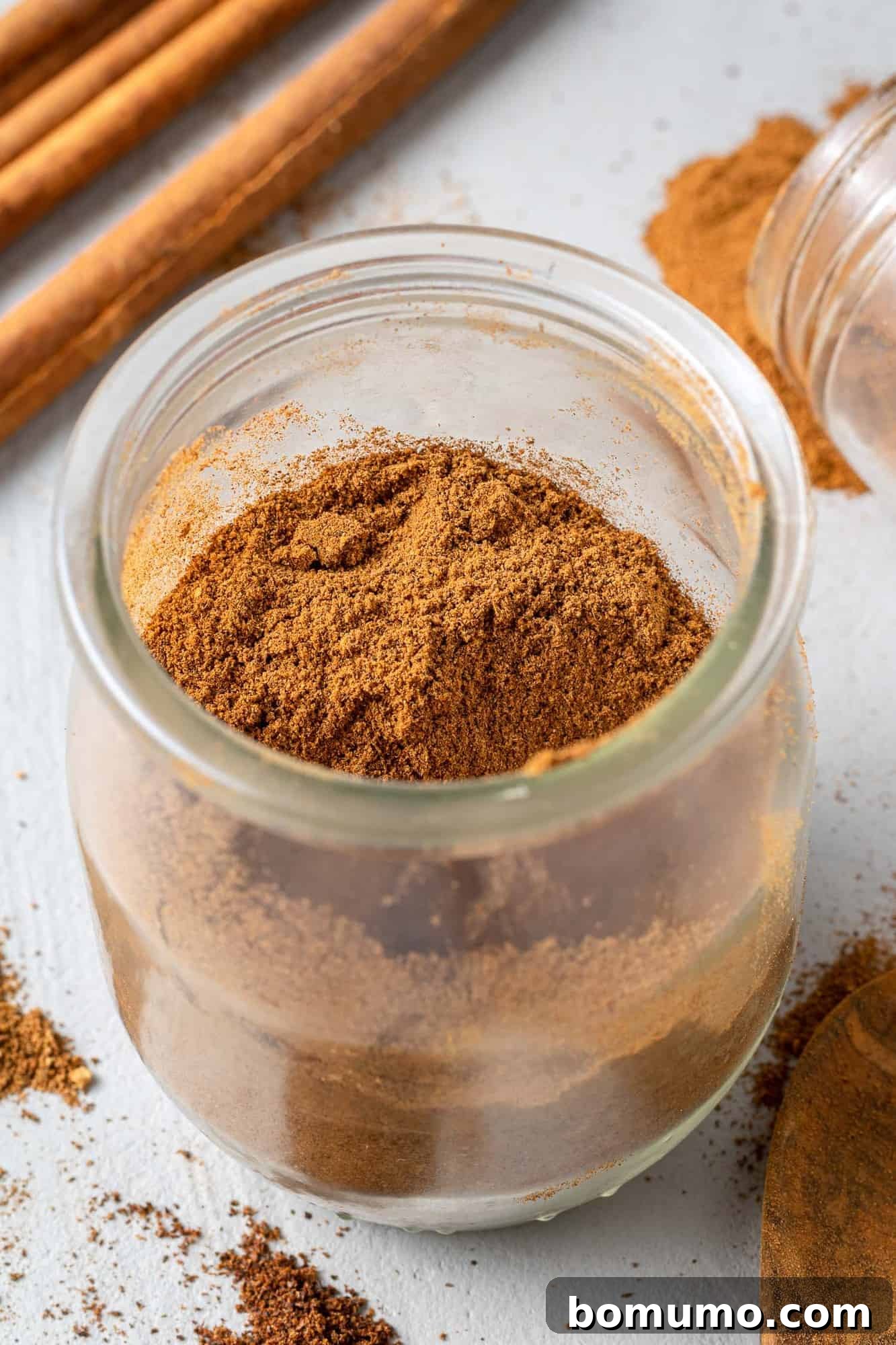 A small glass jar filled with golden-brown homemade pumpkin pie spice, adorned with a simple label, sits next to several whole cinnamon sticks, against a rustic wooden background.