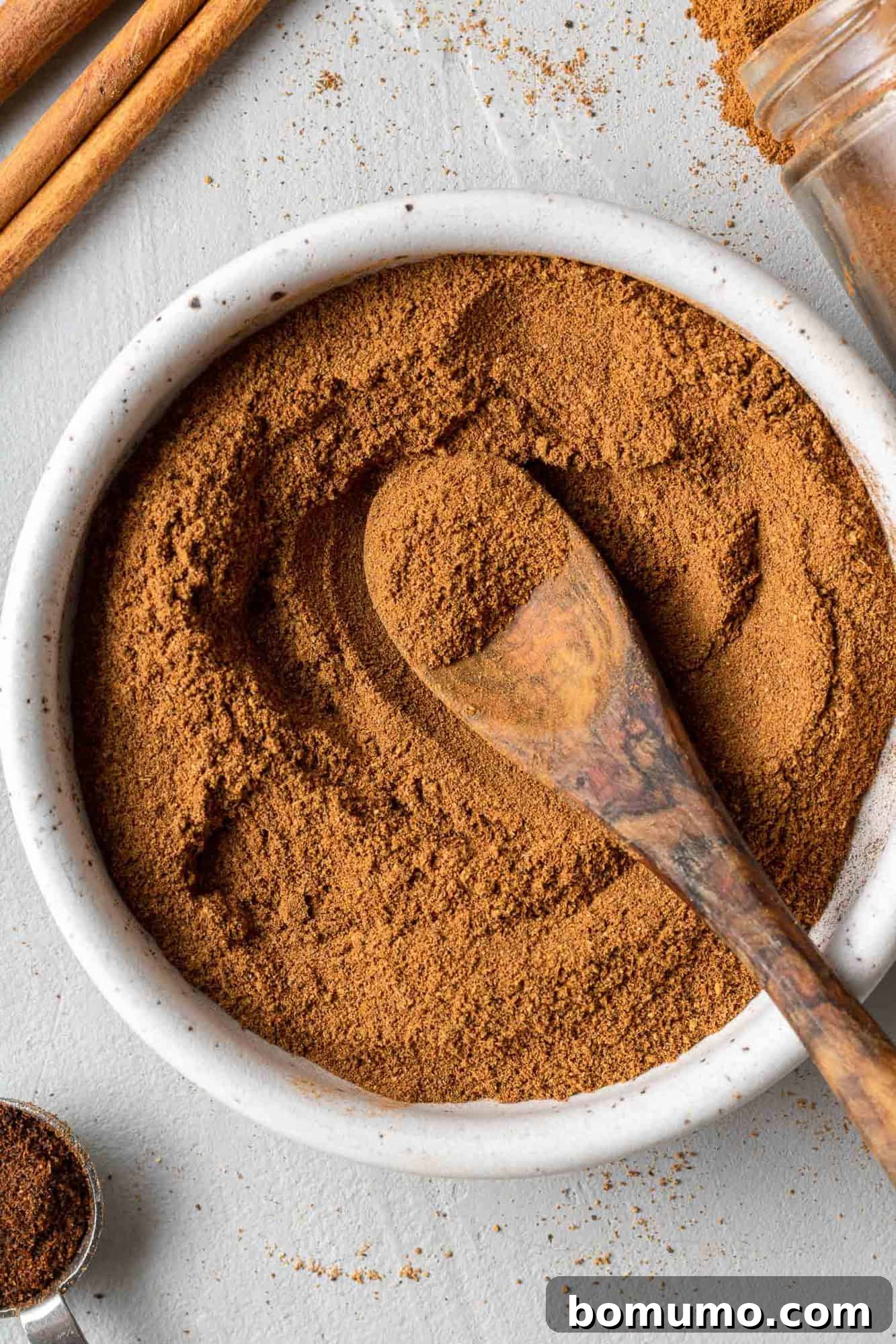 A small white bowl filled with homemade pumpkin pie spice, featuring a tiny wooden spoon resting inside, all against a warm, inviting backdrop.