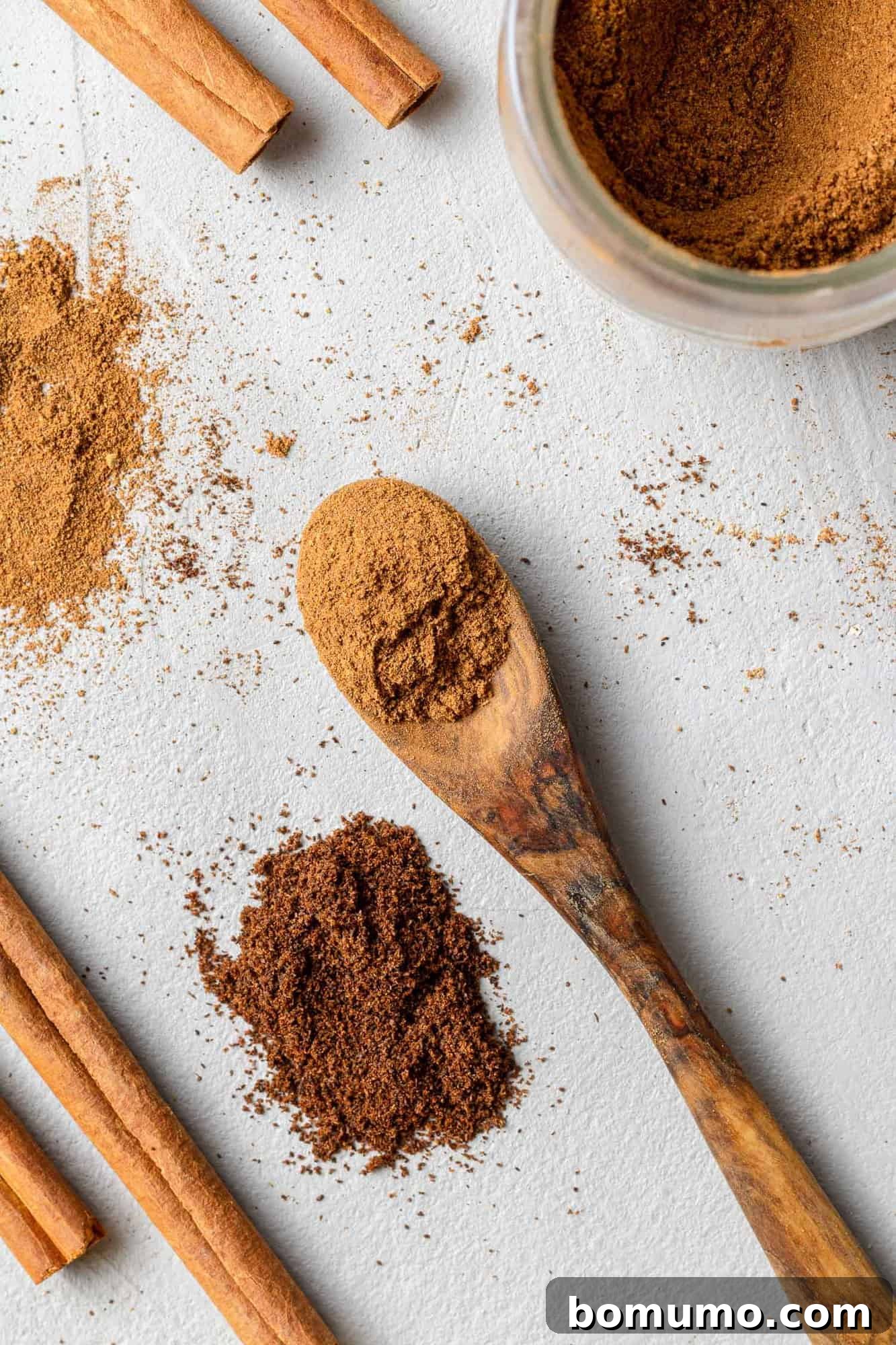 A tiny wooden spoon holds a small mound of finely ground brown spices, with other various spices scattered elegantly nearby on a textured surface.