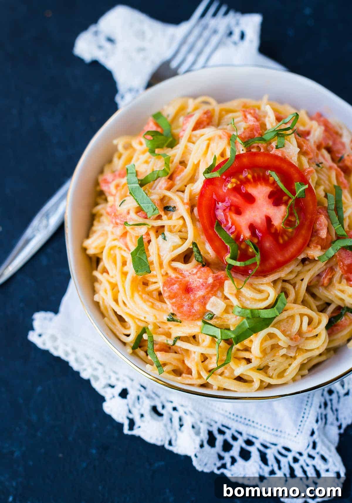 A bowl of angel hair pasta coated in a light cream and fresh tomato sauce, garnished with a slice of fresh red tomato and basil.