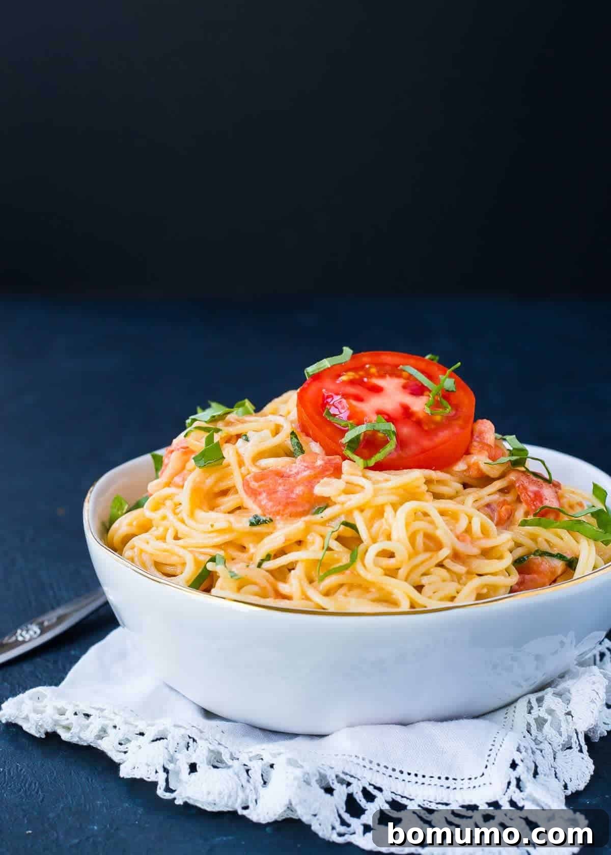 A white bowl filled with angel hair pasta and tomato cream sauce, garnished with a slice of tomato and fresh basil.