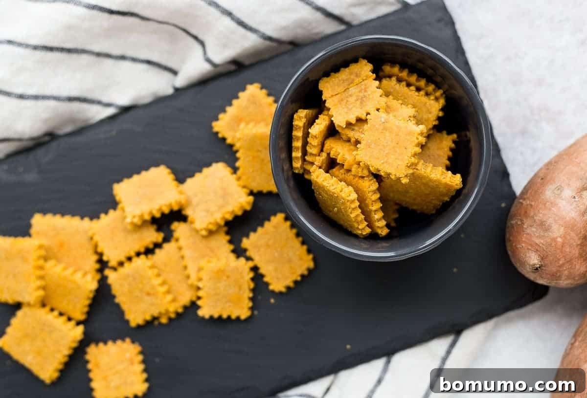 An overhead shot showcasing a rustic bowl filled with golden sweet potato and rosemary gluten-free crackers, with more scattered across a dark slate surface, emphasizing their crispy texture and inviting presentation.