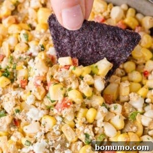 A blue tortilla chip is dipped into a mixture of corn, diced red and green onions, and crumbled cheese.