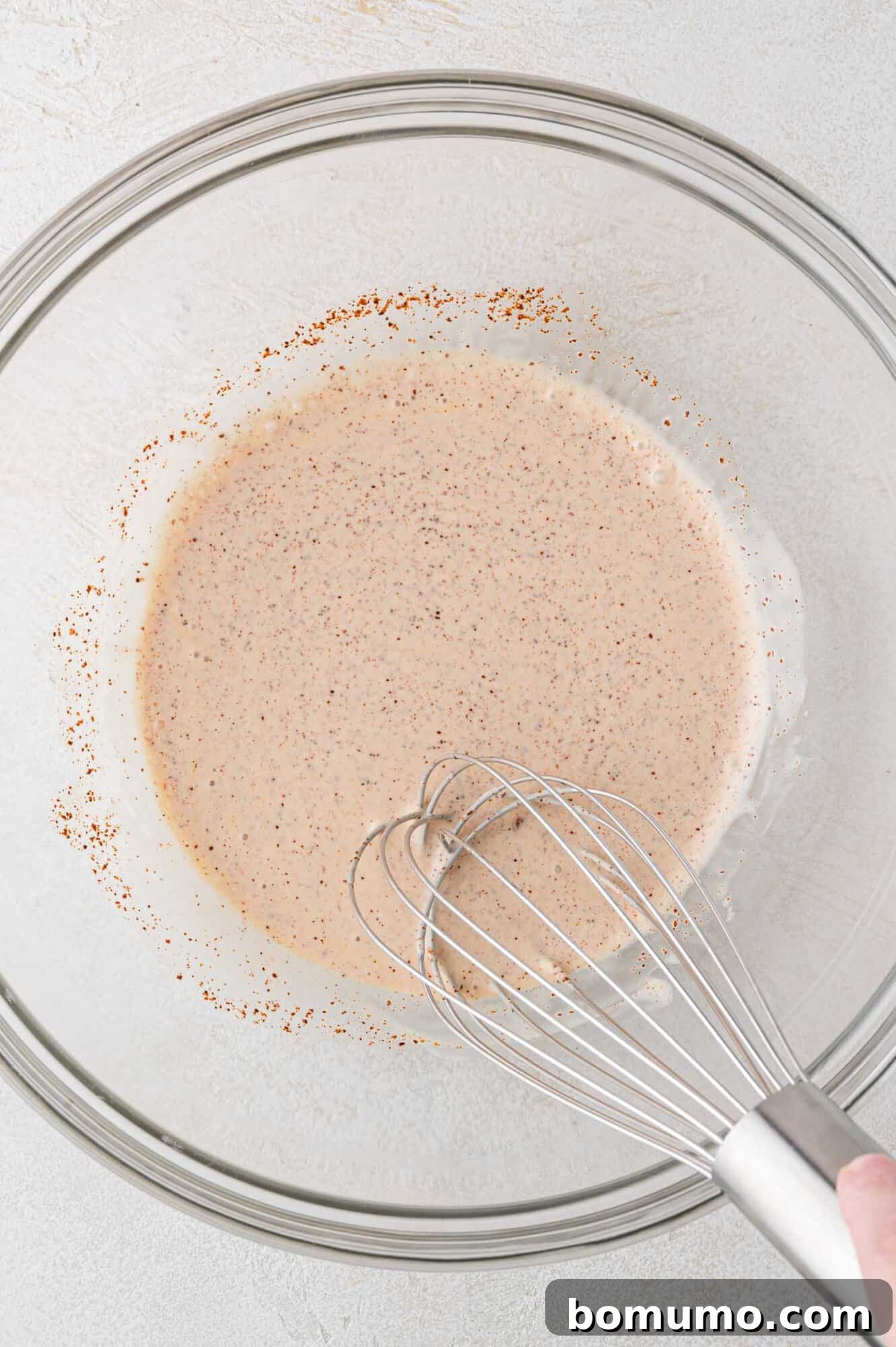 A whisk stirring a creamy, speckled mixture in a clear glass bowl on a light-colored surface, demonstrating the mixing of the dip's base.