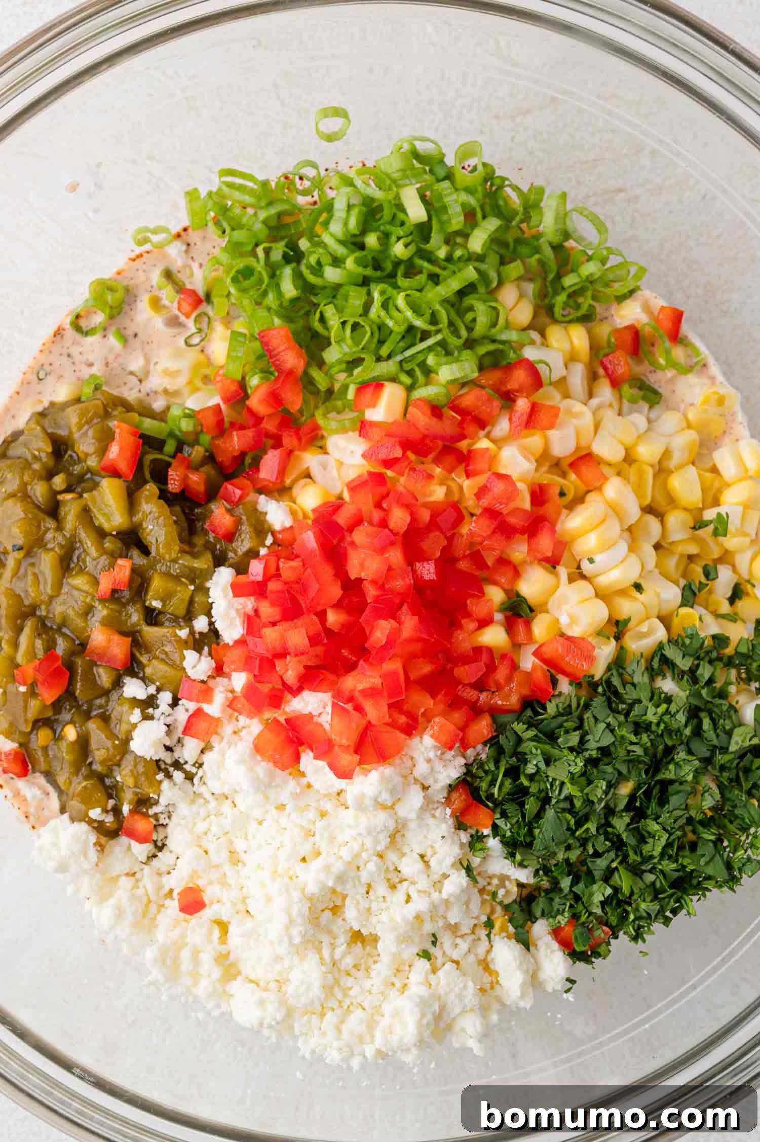 A bowl with chopped red peppers, corn, diced green chiles, scallions, parsley, and crumbled cheese arranged in sections, ready to be combined into the dip.
