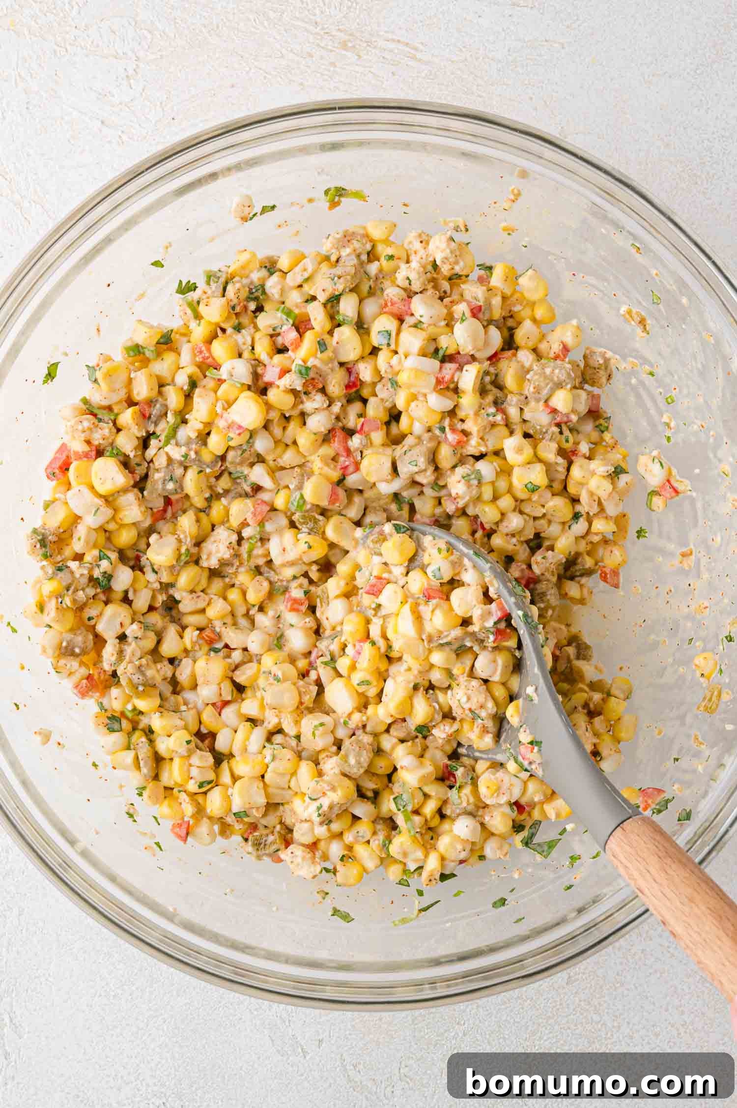 A glass bowl filled with a corn dip being stirred with a large serving spoon, showing the final mixture of ingredients.