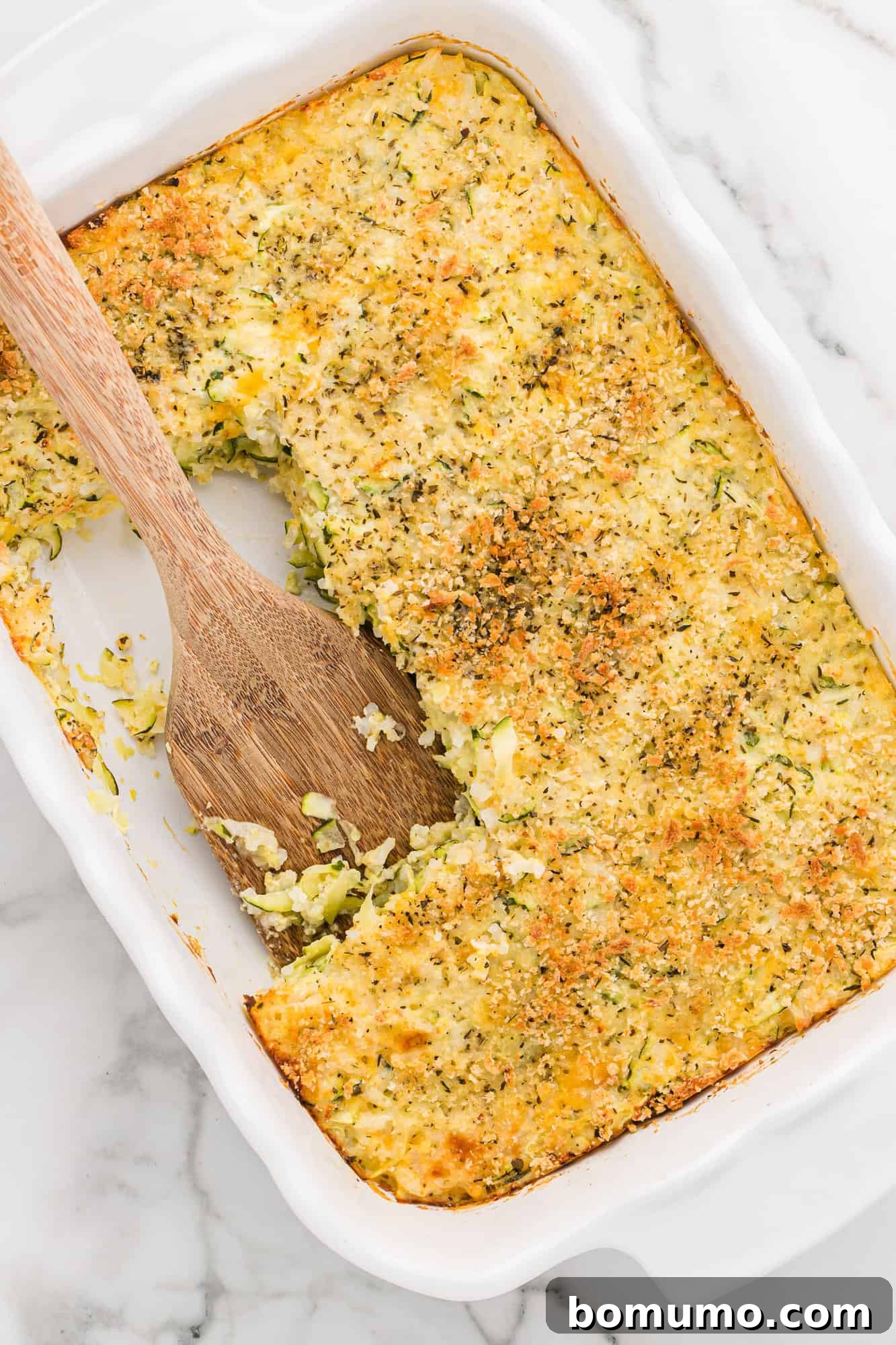 A baking dish of golden-brown zucchini rice casserole with a few squares scooped out, showing the creamy, cheesy interior.
