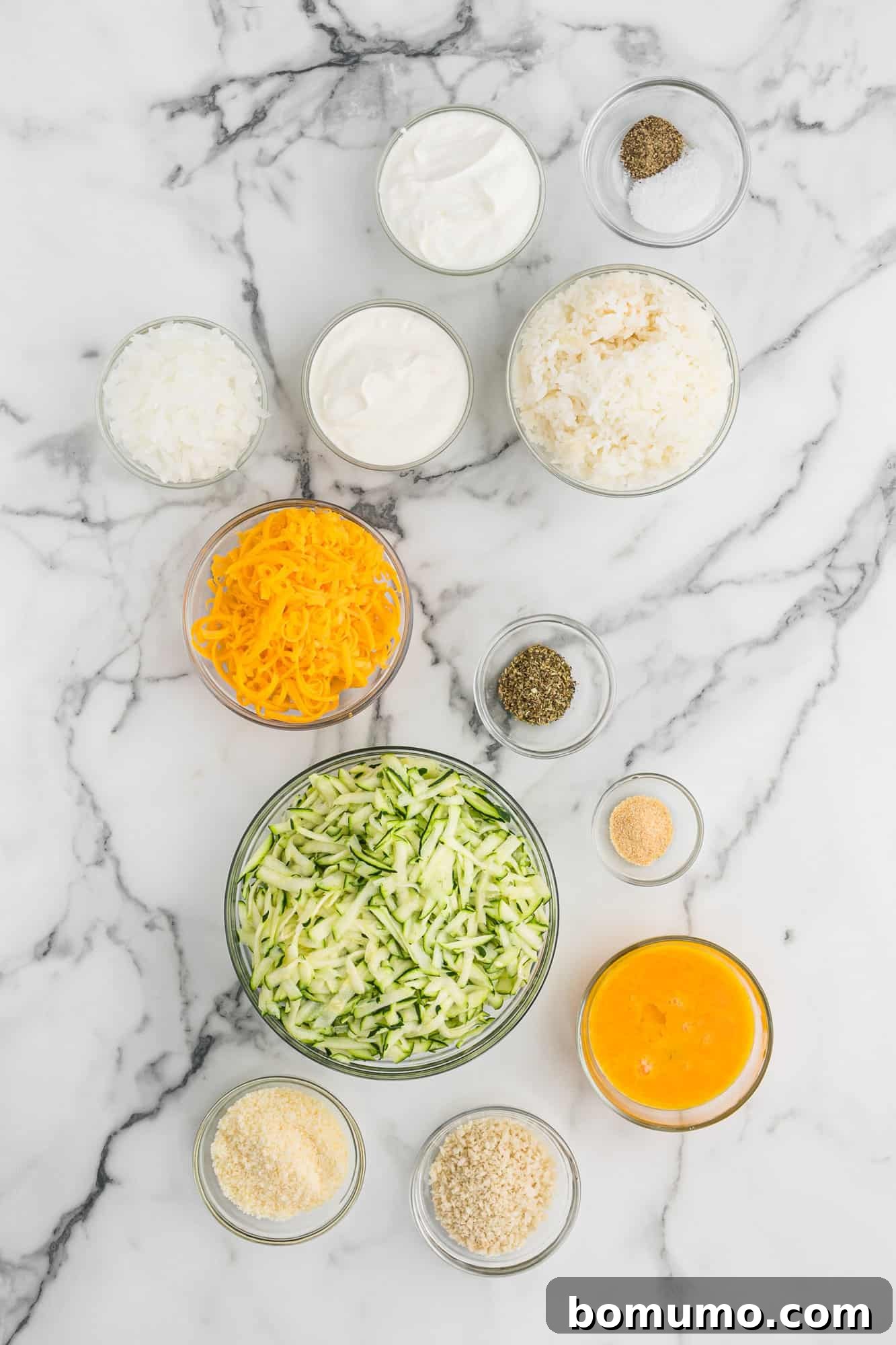 A collection of Zucchini Casserole ingredients including freshly shredded zucchini, blocks of cheese, eggs, sour cream, and spices, ready for mixing.