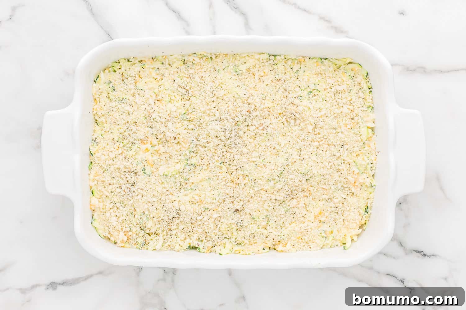 The unbaked zucchini casserole in a baking dish, with the golden panko topping sprinkled evenly on top, ready to go into the oven.