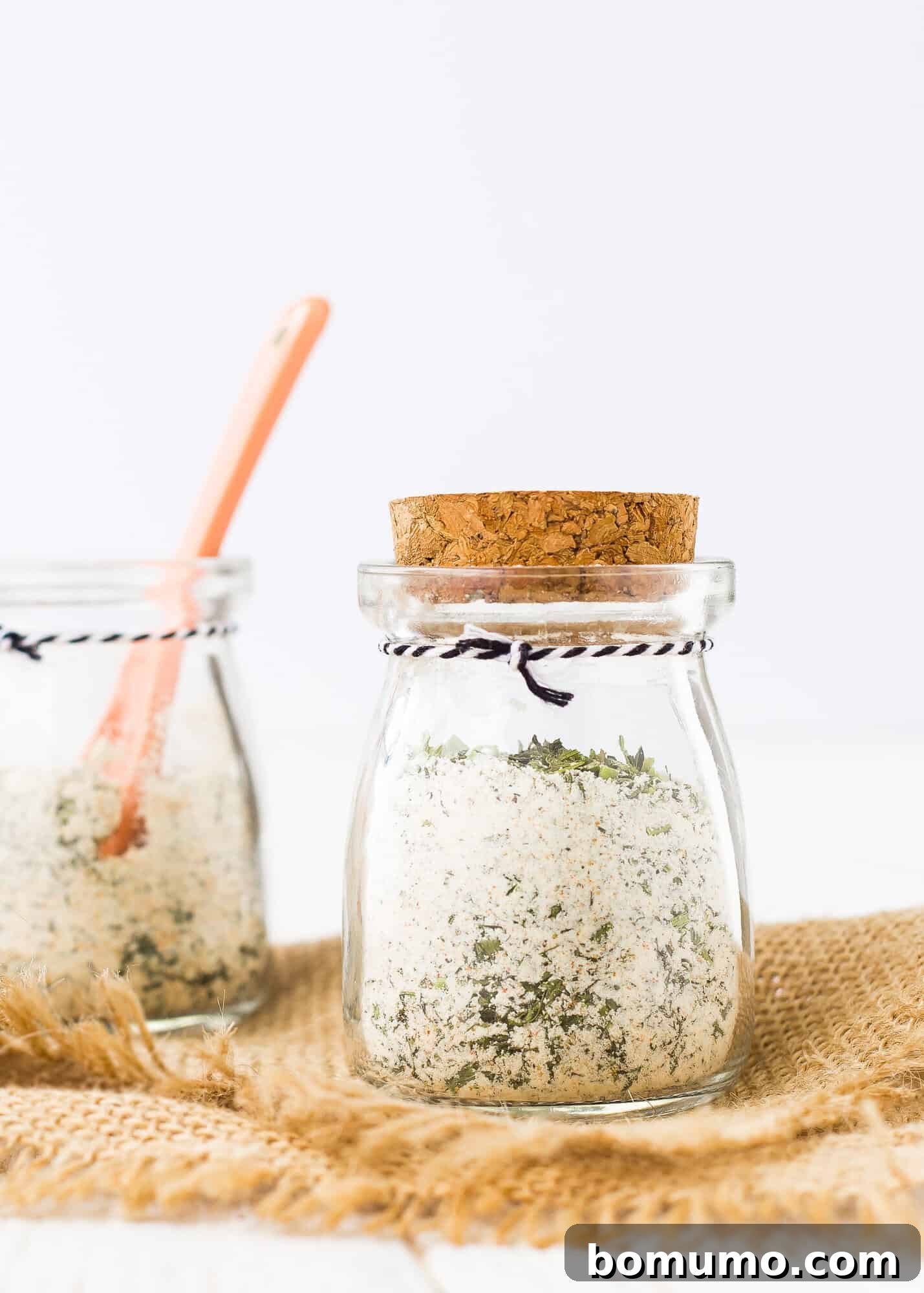 A small, elegant jar filled with homemade buttermilk ranch seasoning mix, with more seasoning in the background, ready for use.